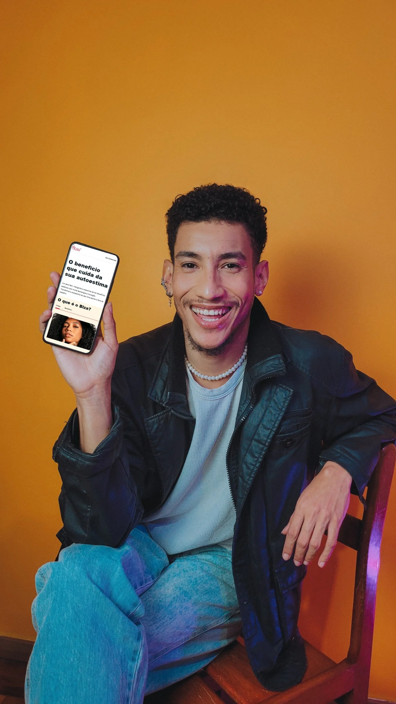 Jovem sorridente sentado em uma cadeira de madeira, segurando um celular com uma apresentação sobre autoestima, com fundo laranja.