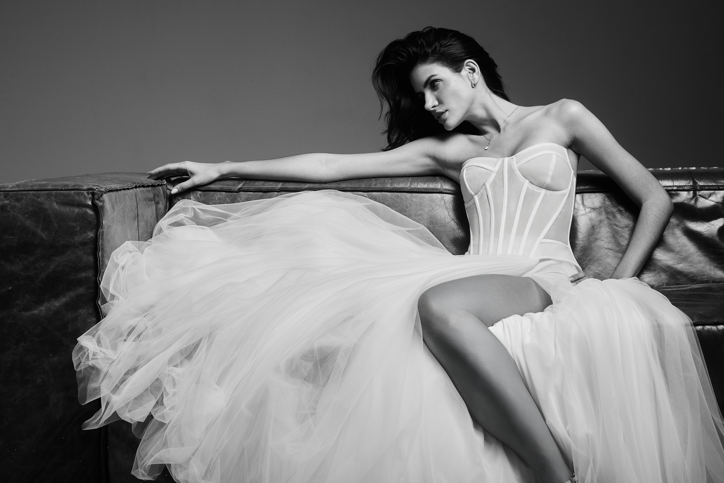 Mulher com cabelo escuro e vestido branco de noiva, deitada em um sofá de couro, em imagem preto e branco.