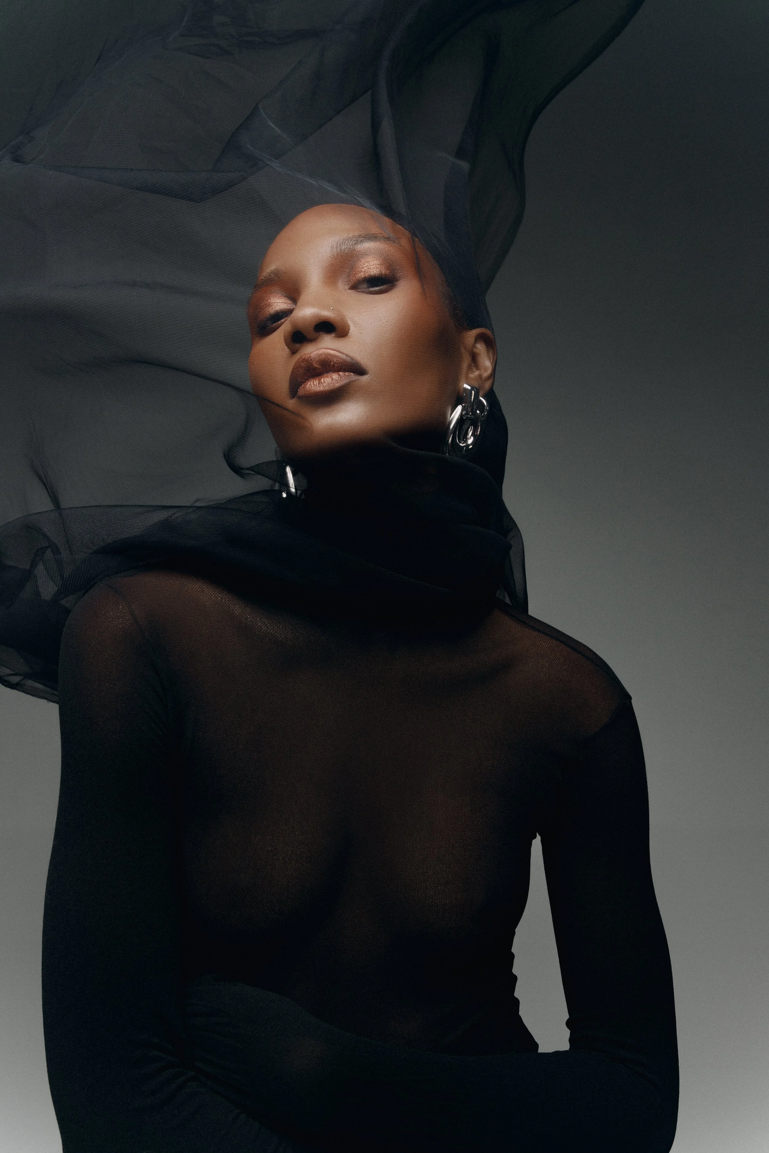 Retrato de uma mulher negra com maquiagem sofisticada, usando roupas pretas transparentes e um acessório de orelha grande. Ela está encarando a câmera com expressão confiante, com fundo cinza escuro.