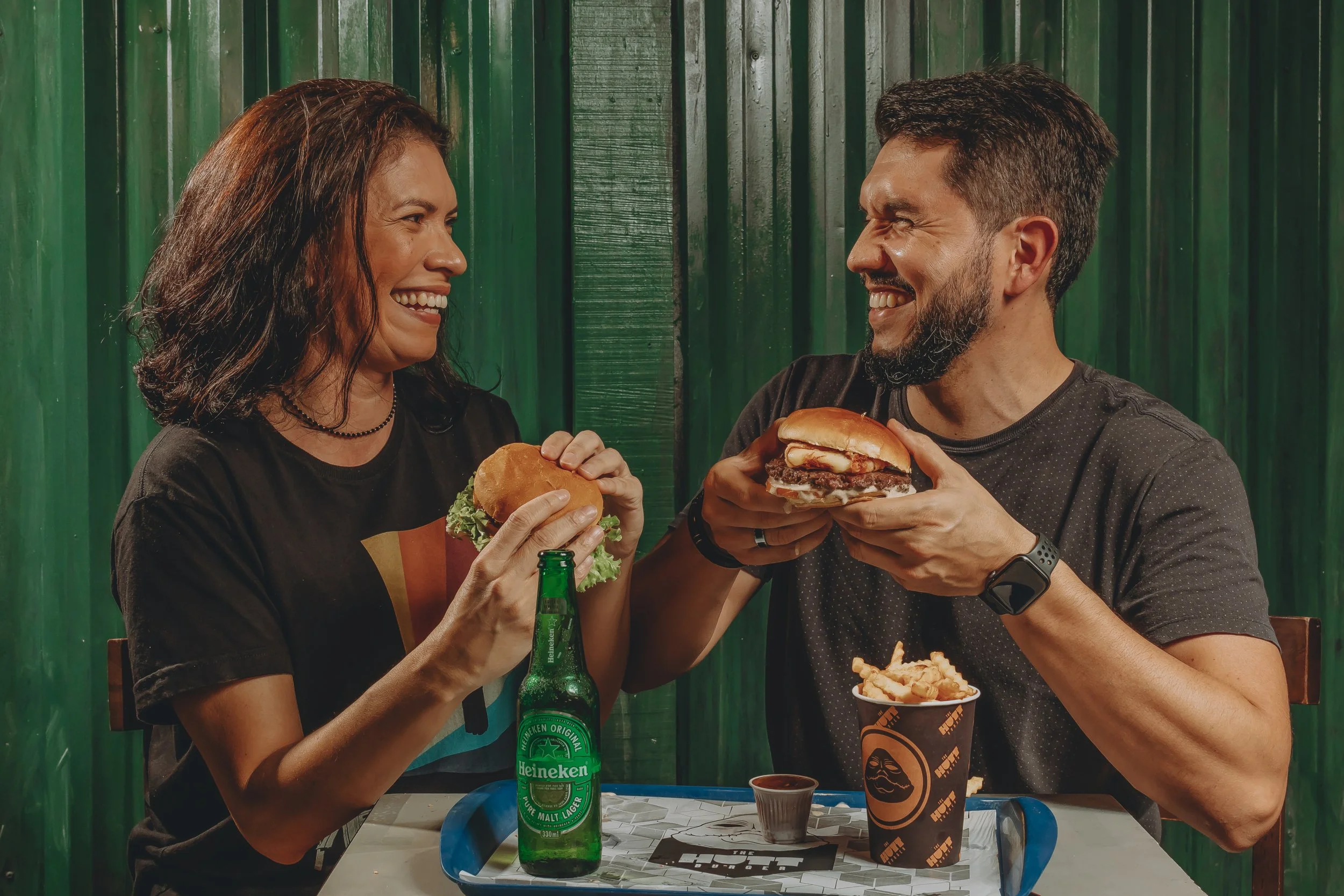 Homens e mulher sorrindo enquanto seguram hambúrgueres em uma mesa com refrigerante e comida, com fundo de parede verde.