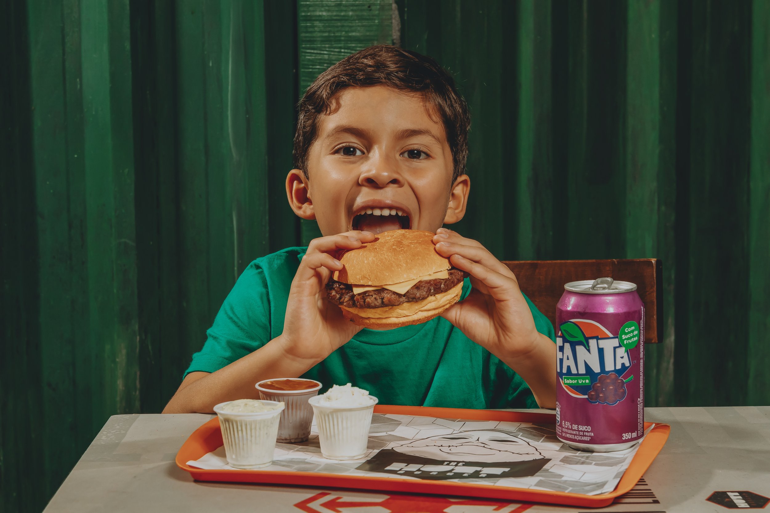 Criança sorridente segurando um hambúrguer na frente da mesa com acompanhamentos e uma lata de refrigerante, sentado em uma cadeira de madeira, com fundo verde