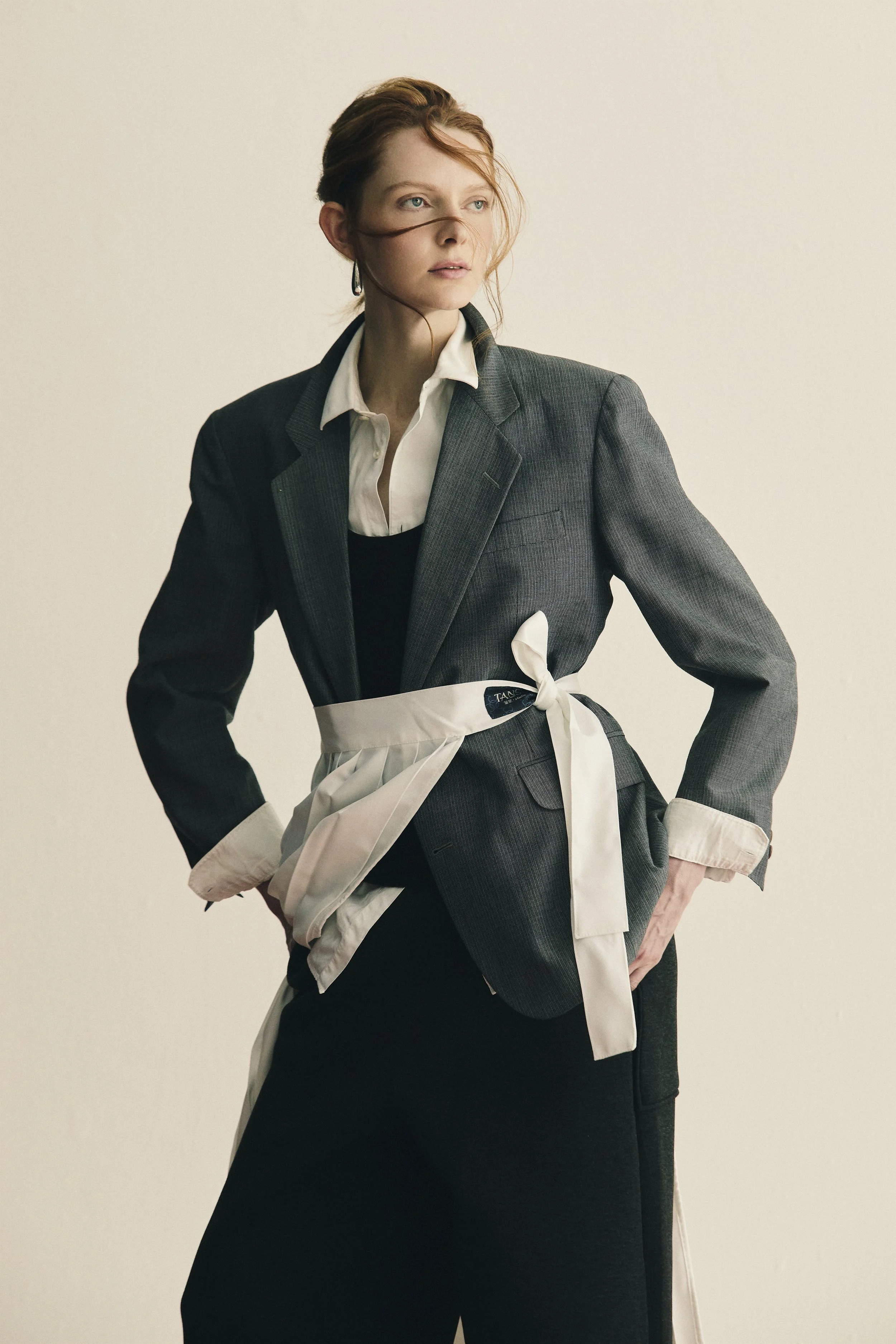 Mulher de cabelos castanhos em um blazer cinza, branca com uma saia preta, usando avental branco na cintura, posando de lado com expressão pensativa contra fundo neutro.
