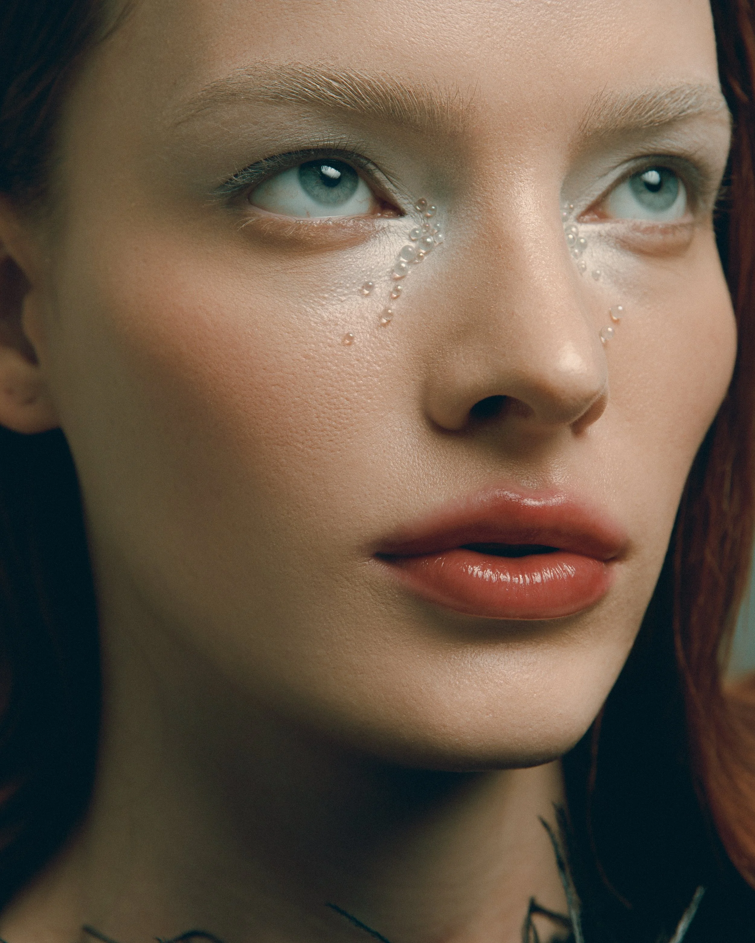 Close-up de rosto de uma mulher com olhos claros e sobrancelhas bem definidas, com pequenas pérolas decorativas sob os olhos.