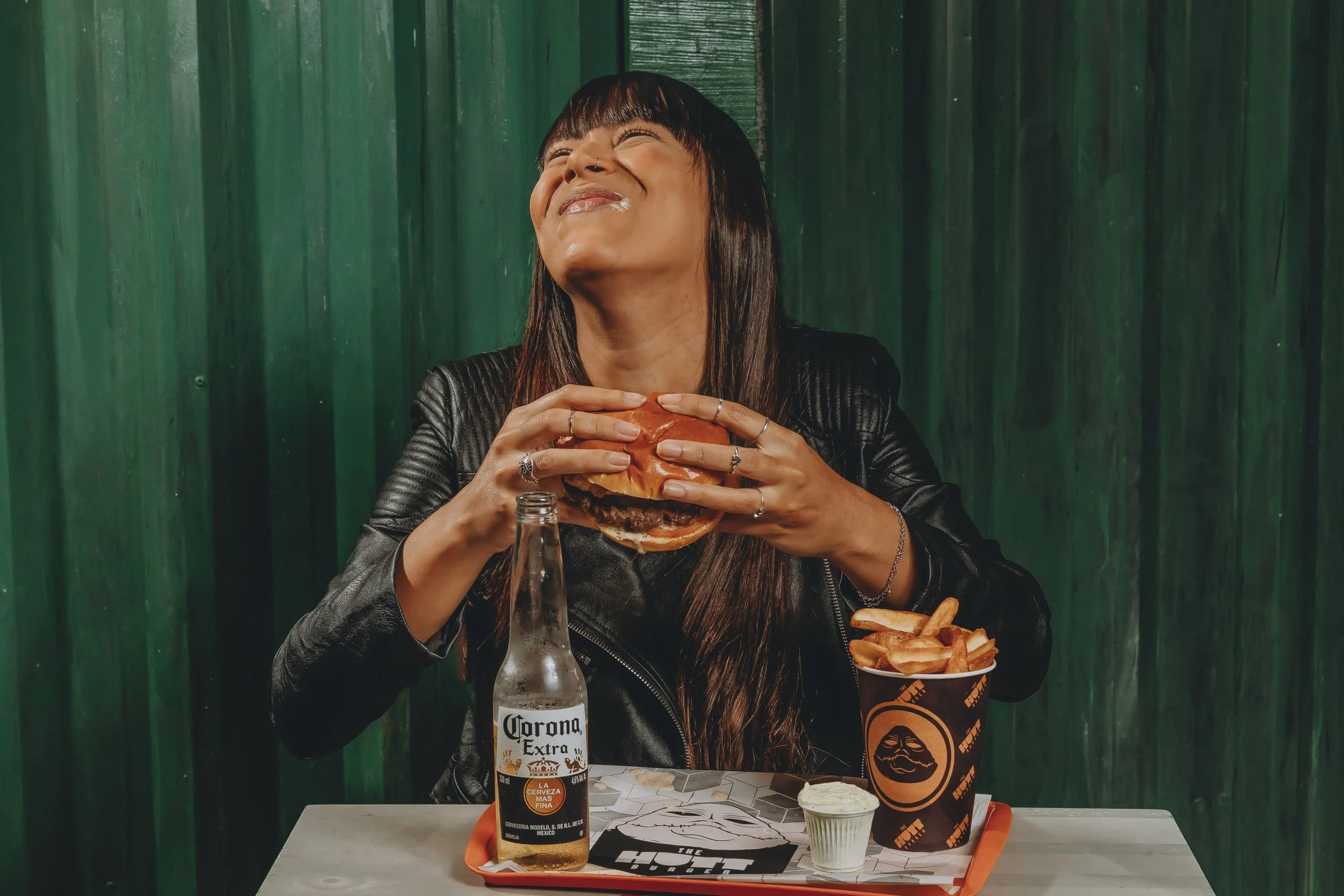 Pessoa com cabelo liso escuro e franja, usando jaqueta de couro preta, sorrindo com olhos fechados, segurando um hambúrguer em um restaurante com parede de madeira verde ao fundo. Na mesa, há uma garrafa de cerveja Corona, um copo de refrigerante, batatas fritas em um balde de papel e um copo de papel com logo de restaurante.