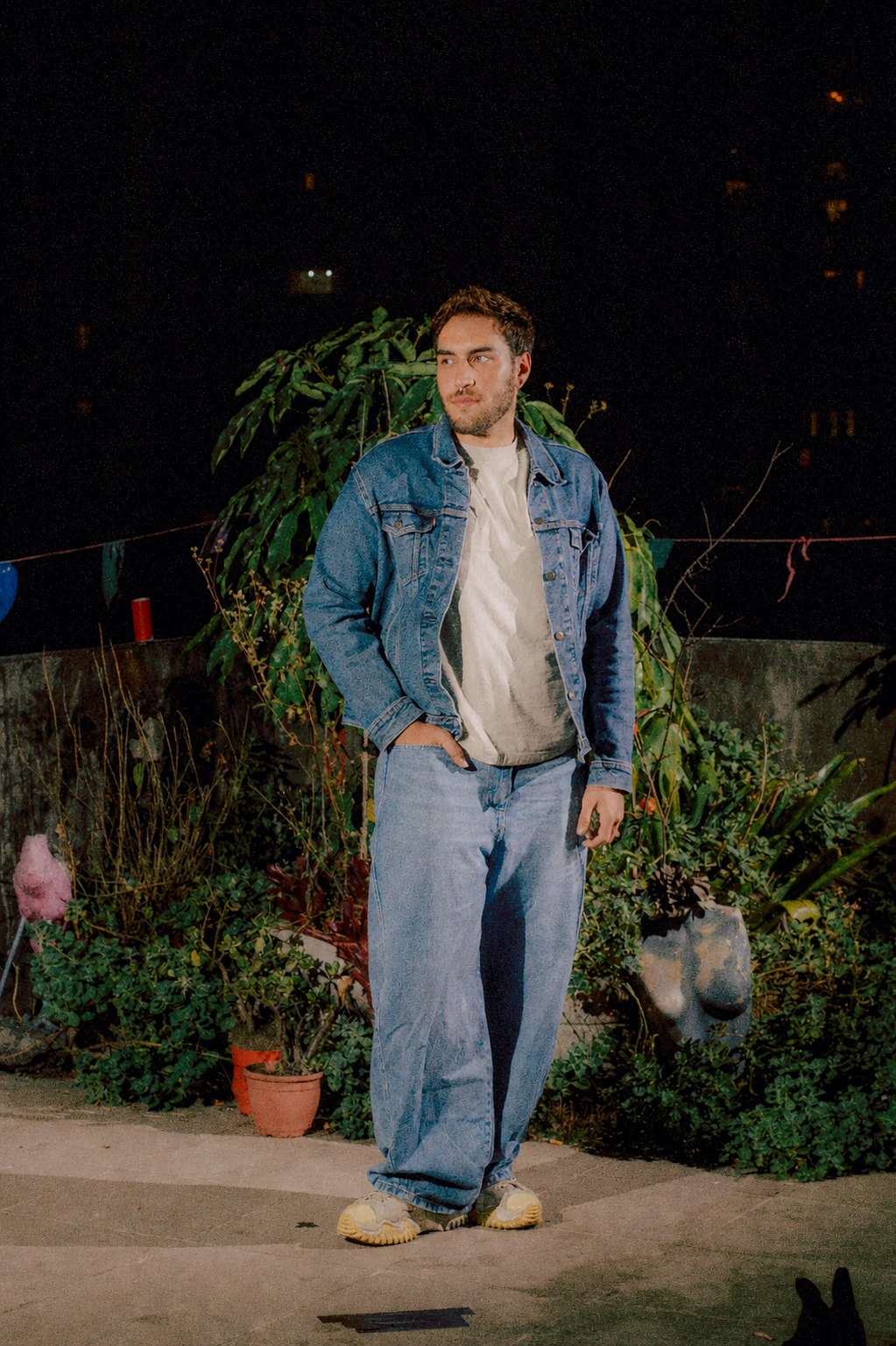 Homem jovem de cabelo castanho, barba curta, usando jaqueta jeans, camiseta branca e jeans largo, segurando um celular na mão esquerda, posando ao lado de plantas e objetos decorativos ao ar livre à noite.