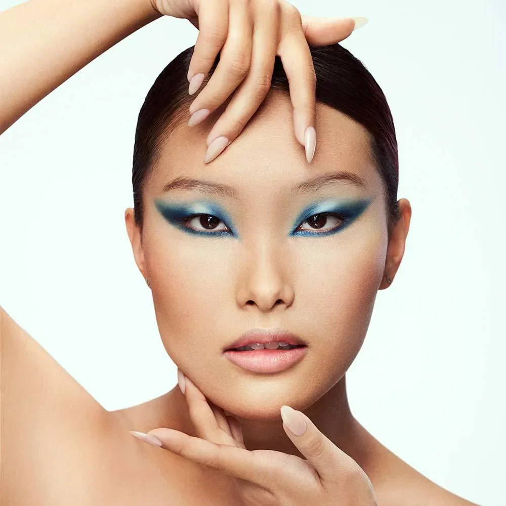 Mulher com maquiagem chamativa, destaque para olhos com sombra azul escura e delineado marcante, cabelo escuro e pele clara, posando com uma expressão séria.