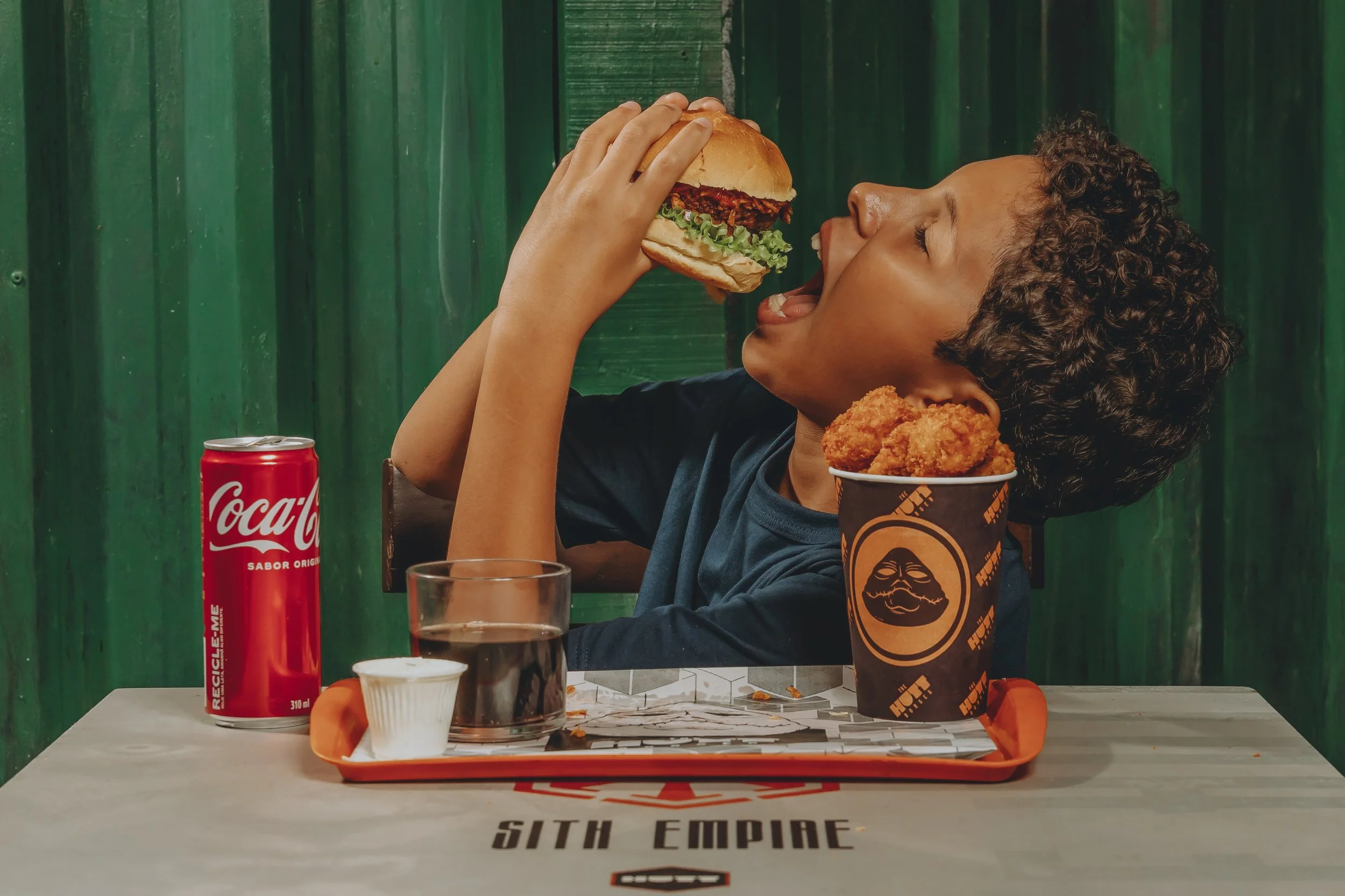 Menina sorridente segurando um hambúrguer próximo ao rosto, sentado em uma mesa com uma bandeja de fast food que inclui um balde de frango frito, duas bebidas, e um prato pequeno com queijo derretido. Fundo com parede verde.
