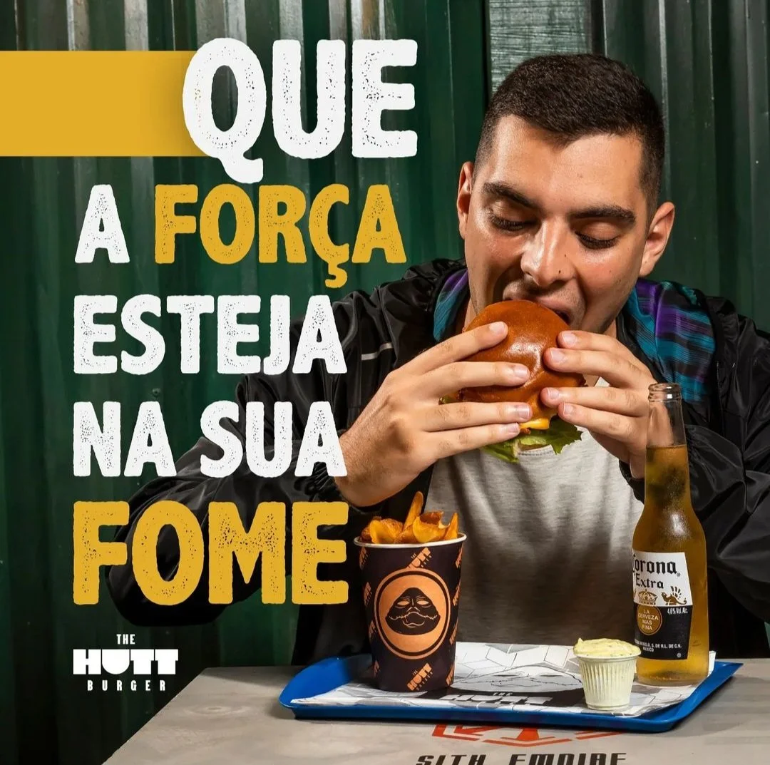 Homem comendo um hambúrguer em um restaurante de fast food, com uma cerveja e batatas fritas na mesa, ao lado de um recipiente de molho e uma porção de manteiga, com fundo de paredes verdes e uma mensagem em português.