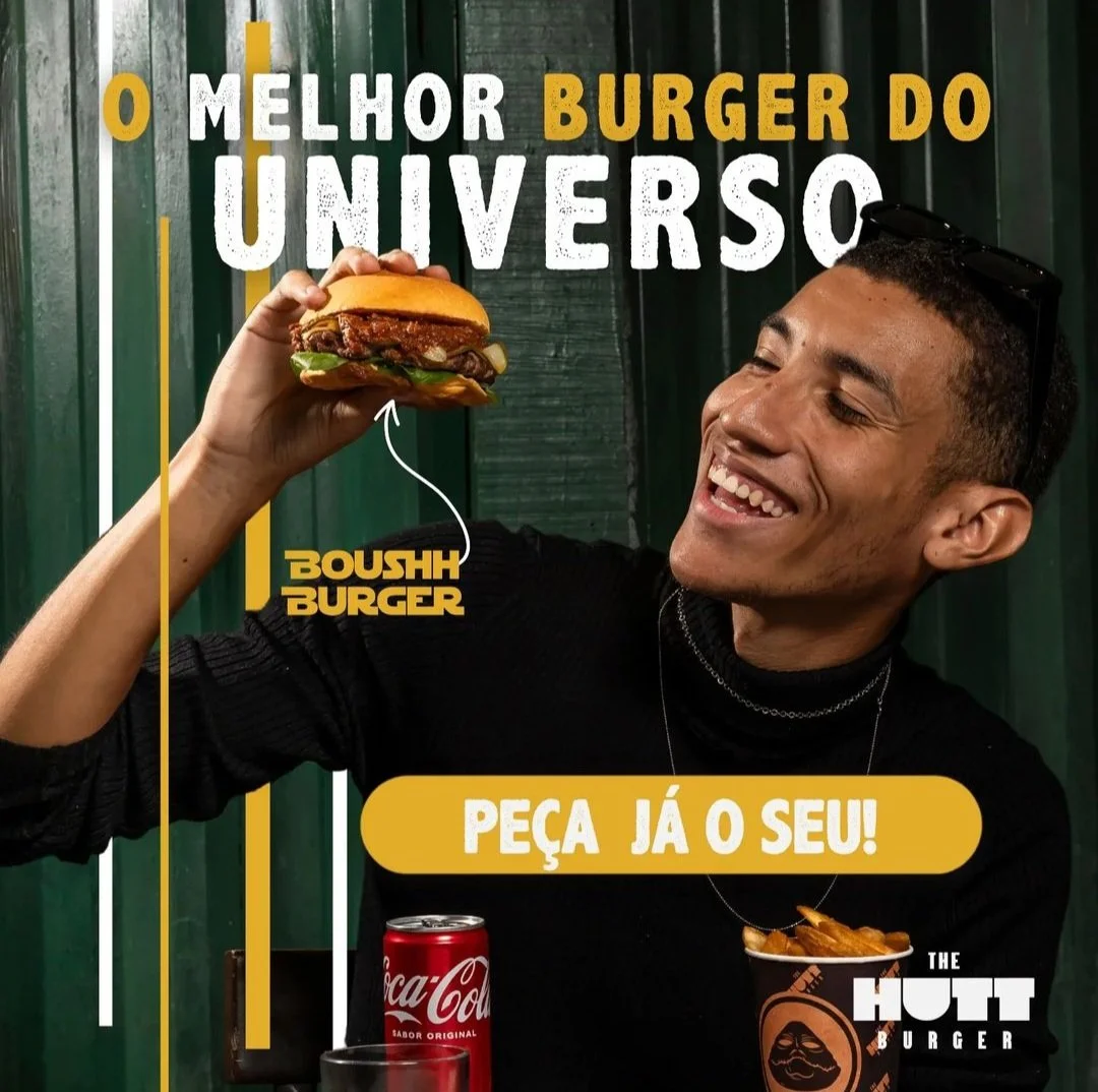 Homem sorridente segurando um hambúrguer no restaurante The Hutt Burger, com uma latinha de Coca-Cola ao lado, parede verde ao fundo, promovendo o melhor hambúrguer do universo.