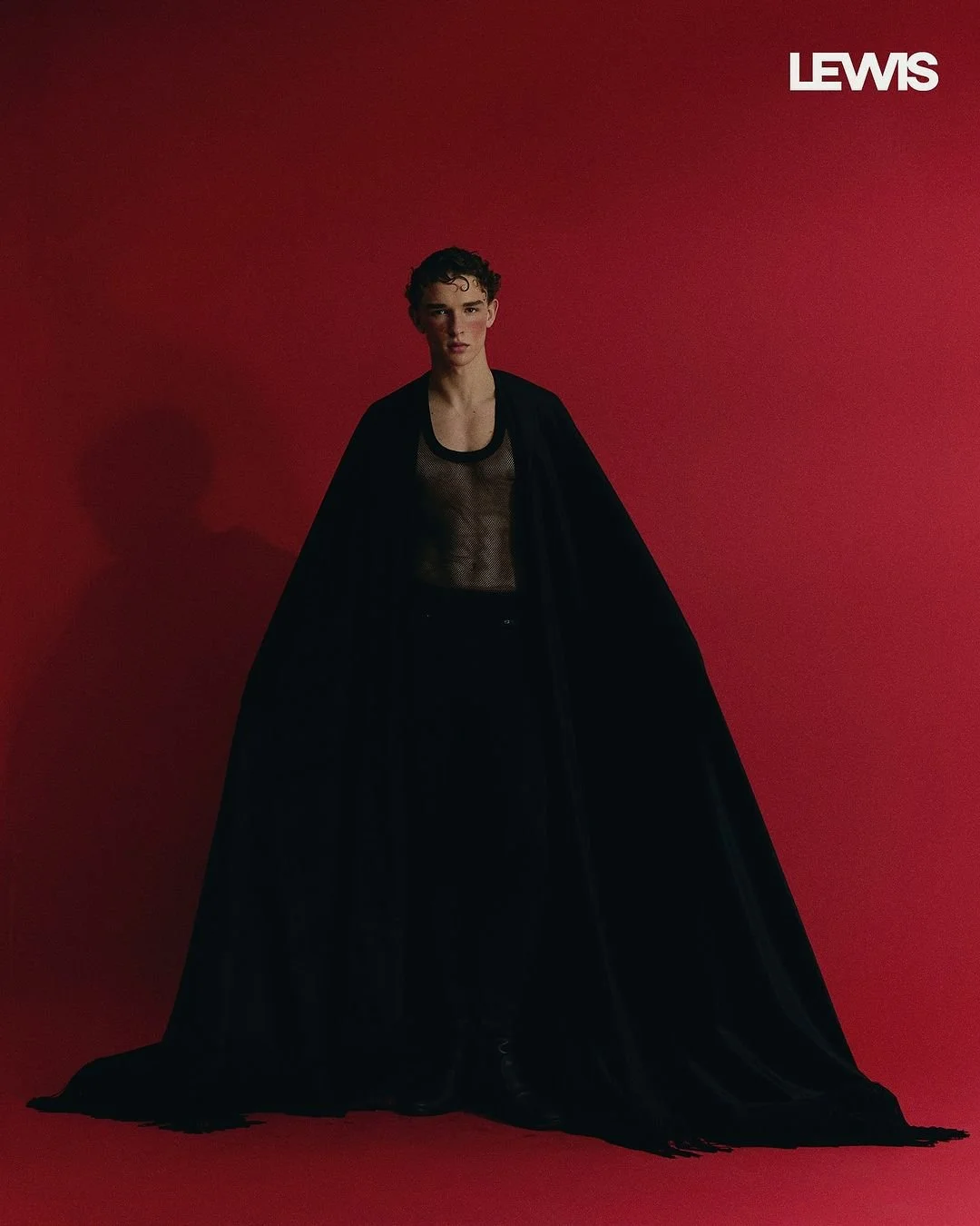 Pessoa jovem com cabelo cacheado, usando roupa preta transparente e capa longa preta, em fundo vermelho