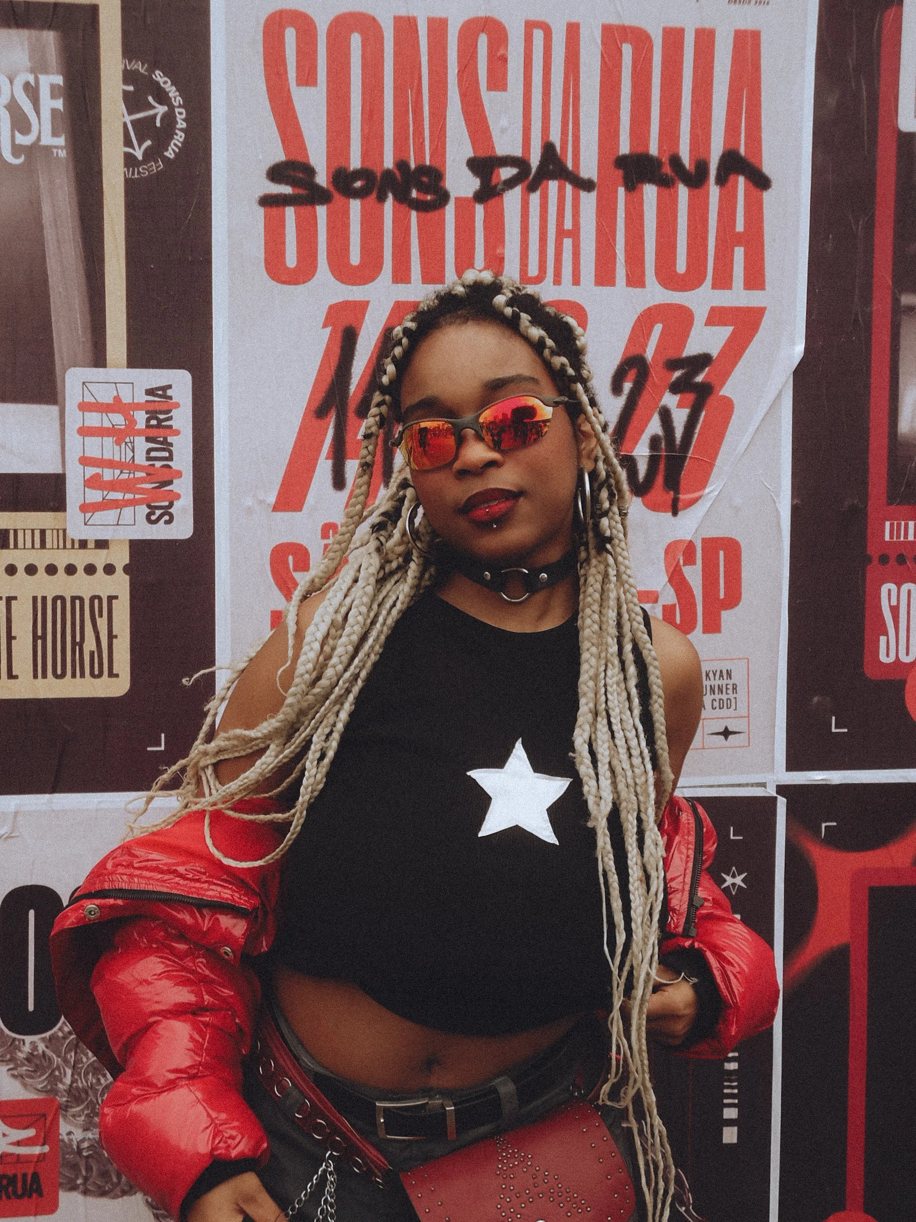 Mulher com cabelo de mechas loiras e pretas usando óculos de espelho vermelho, camiseta preta com uma estrela branca, jaqueta vermelha, calça de couro com detalhes de spikes e brincos grandes, posando diante de um mural com textos em português.