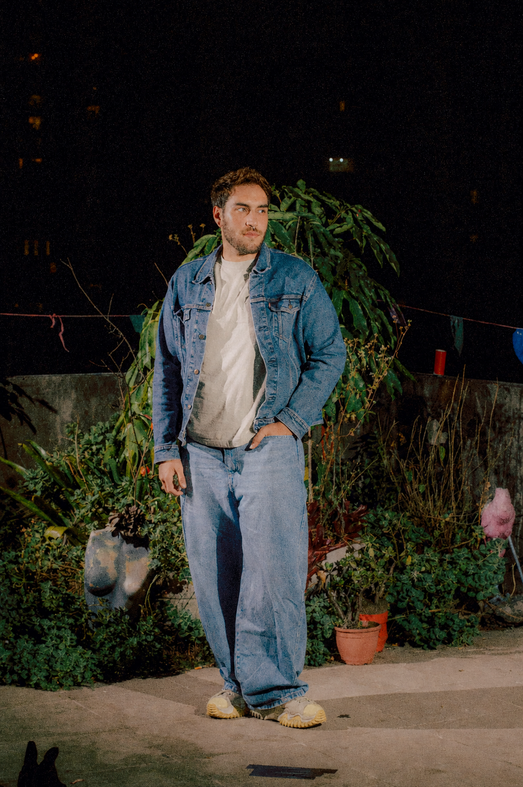 Um homem de cabelo castanho, barba e pele clara usando jaqueta jeans, camiseta cinza e calças largas, de pé em um jardim à noite, ao lado de plantas e uma parede com luzes decorativas.