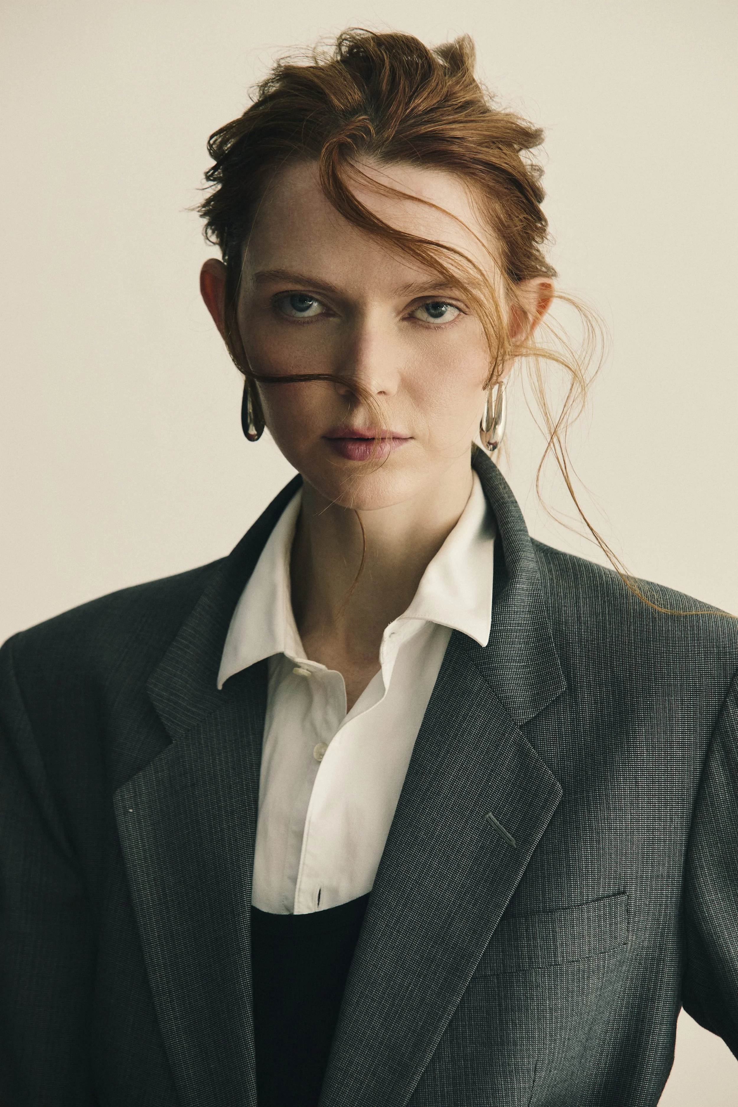 Retrato de uma mulher com cabelo avermelhado e olhos claros, usando terno cinza e camisa branca, com expressão séria, contra fundo claro.