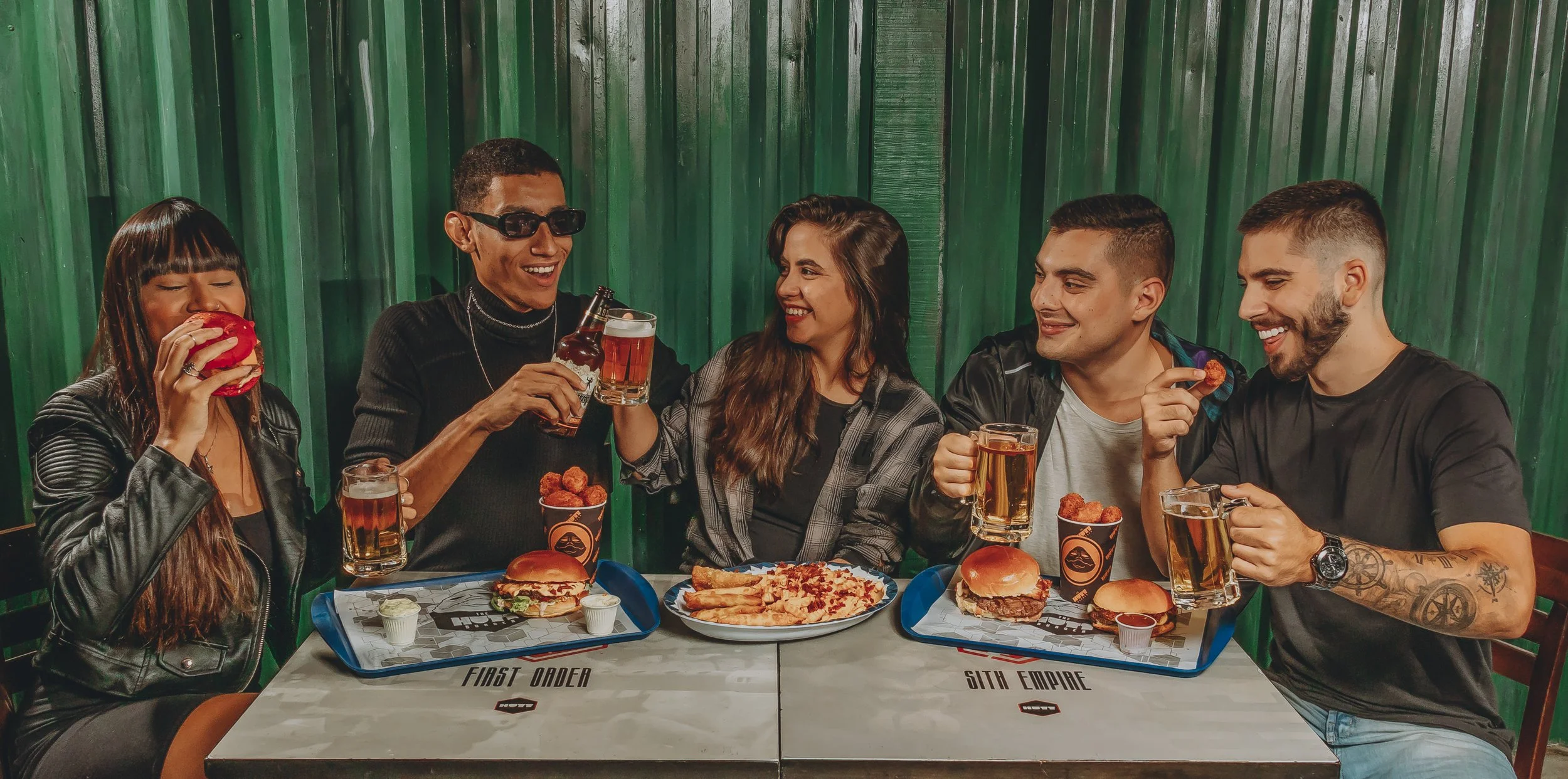 Grupo de seis pessoas sorrindo e brindando com bebidas em um restaurante ou lanchonete, com comida como hambúrgueres, frango frito e pizza na mesa, fundo de parede de metal verde