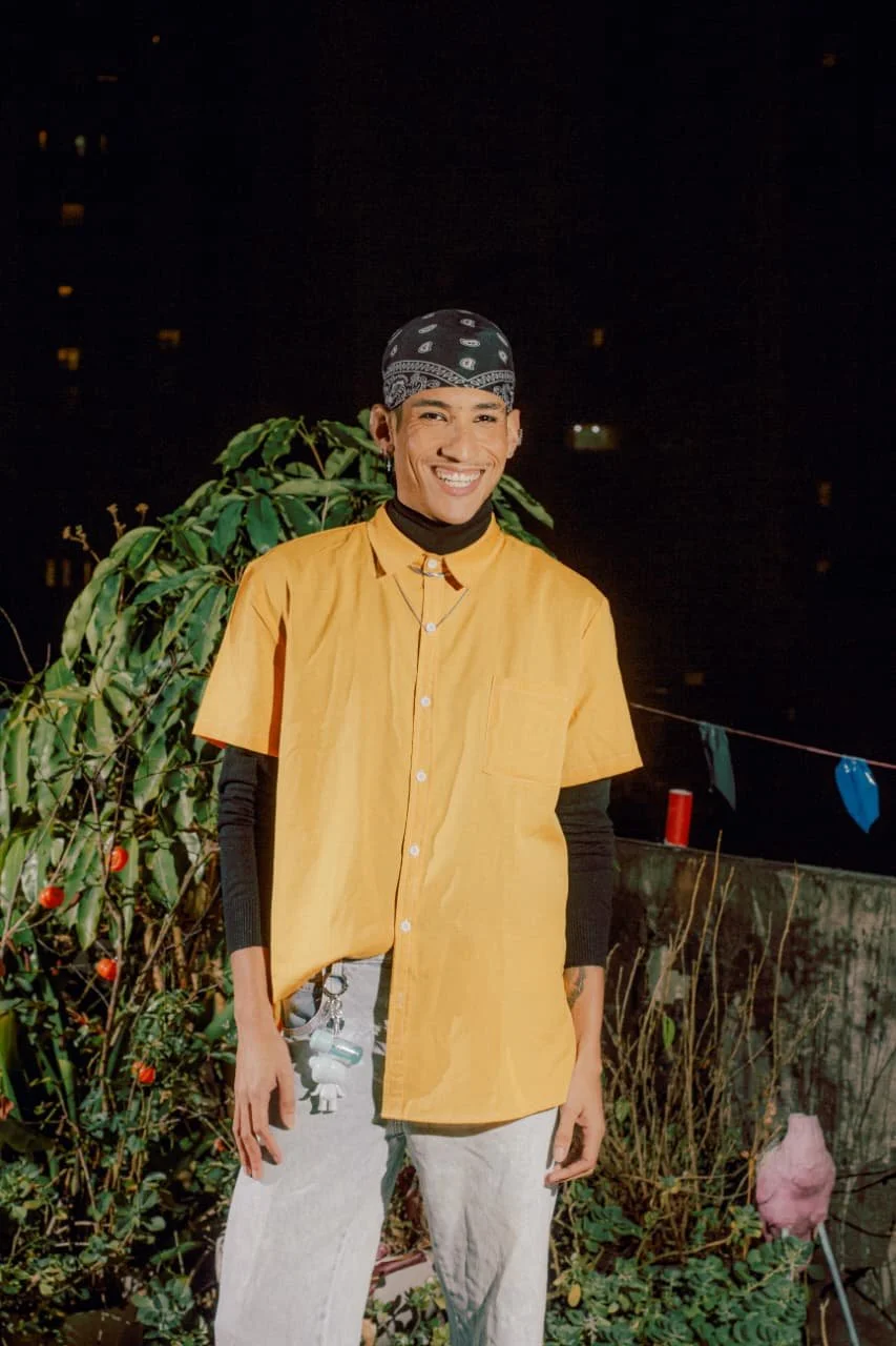 Pessoa sorridente vestindo uma jaqueta amarelo, usando bandana preta com estampa, com plantas ao fundo, durante a noite.