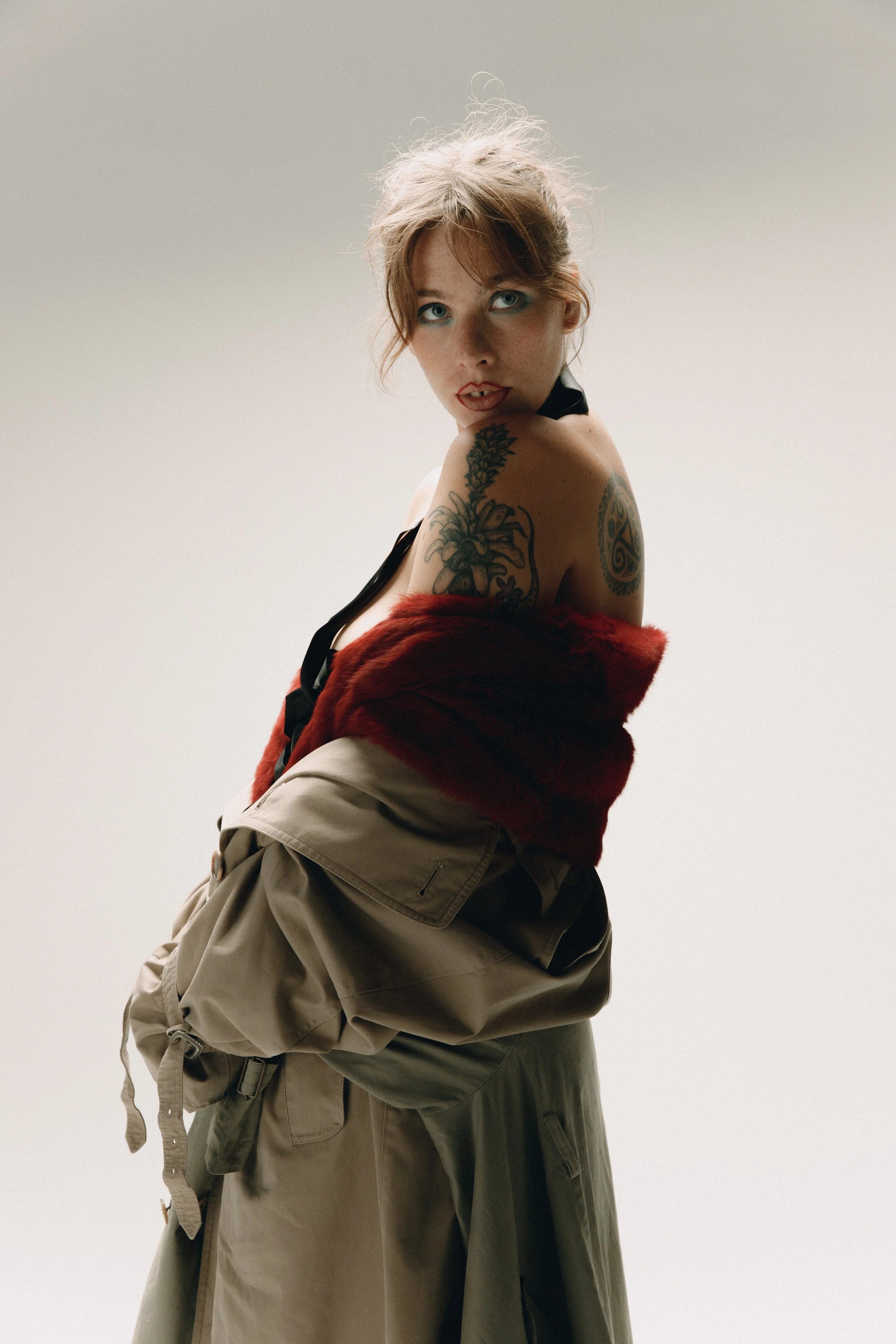 Mulher com cabelos castanhos e olhos claros, usando uma jaqueta vermelha com pele falsa no ombro, uma blusa preta, calça beige, com tatuagens visíveis no braço esquerdo, posando contra fundo claro