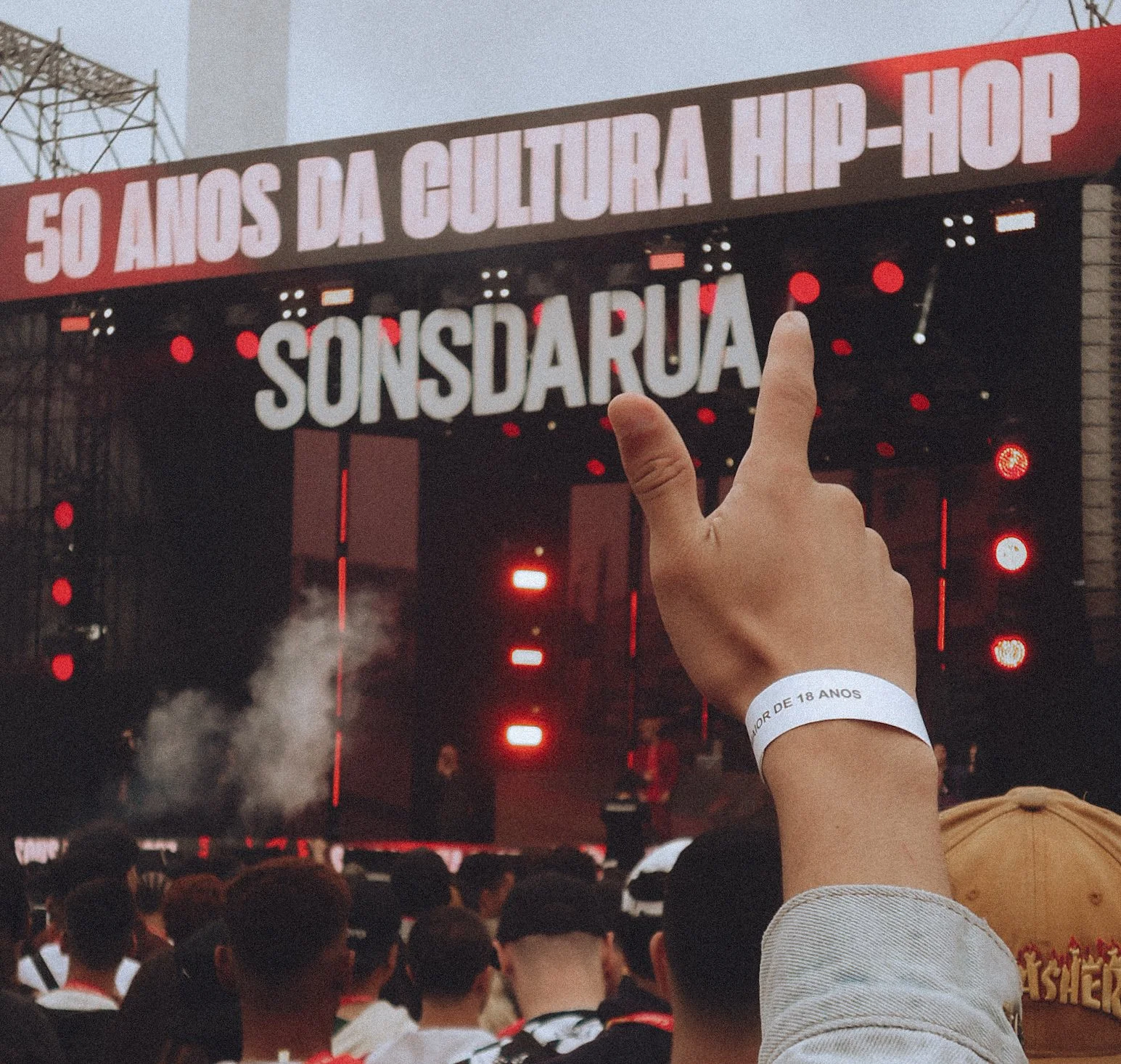 Público assistindo a um evento cultural de hip-hop em uma arena. Uma pessoa levanta o dedo indicador em direção ao palco, que tem uma grande tela com a inscrição "50 ANOS DA CULTURA HIP-HOP" e "SÔNSDA RUA".