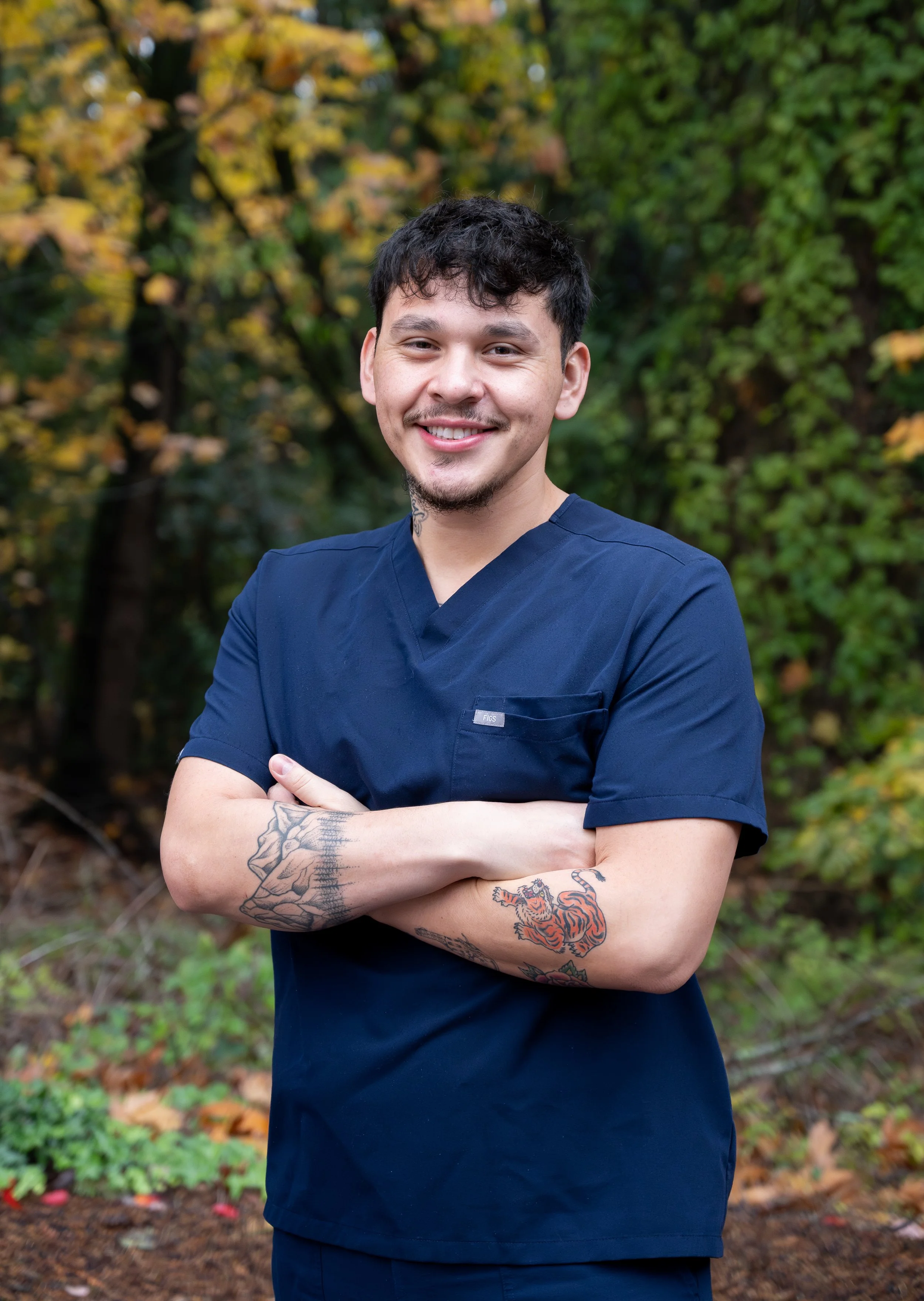 A young man in navy scrubs standing outdoors with arms crossed, smiling, with green trees and autumn leaves in the background.