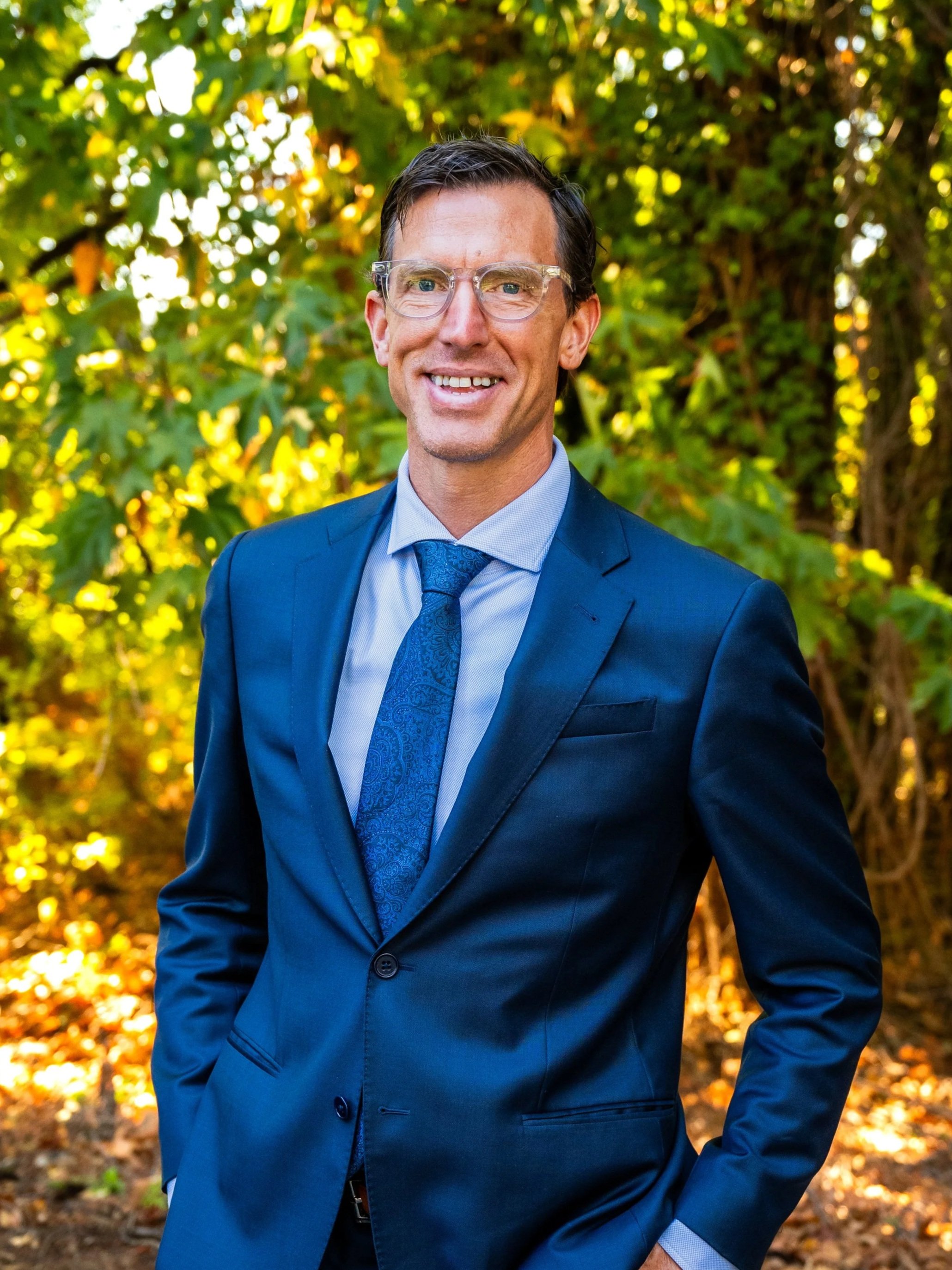 Dr. DeLong in a blue suit and tie standing outdoors with green leaves in the background.