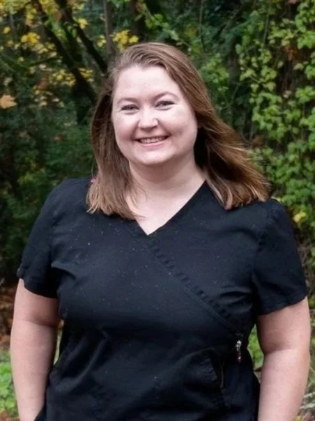 A dental assistant with long reddish-brown hair smiling in front of a blurred green forest background.