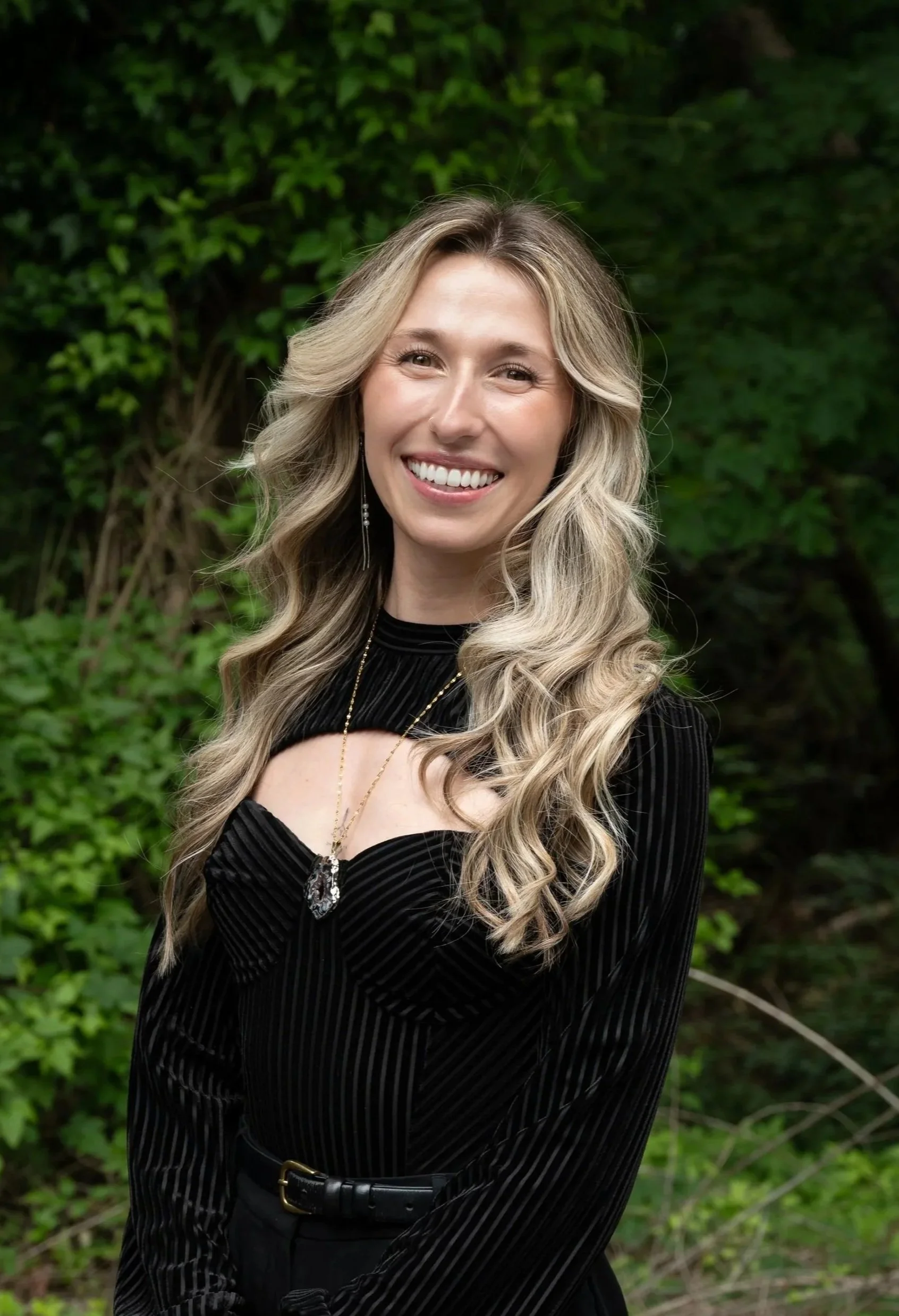 Person smiling outdoors with a background of green foliage.