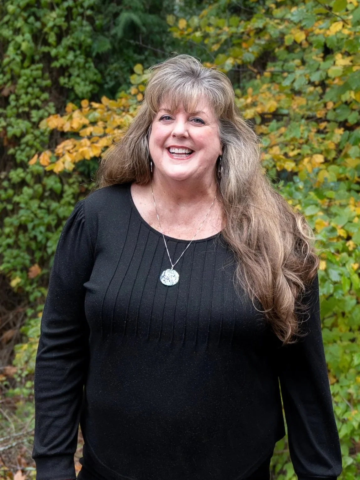 Smiling person with long hair in a green floral top, standing outdoors with trees and greenery in the background.