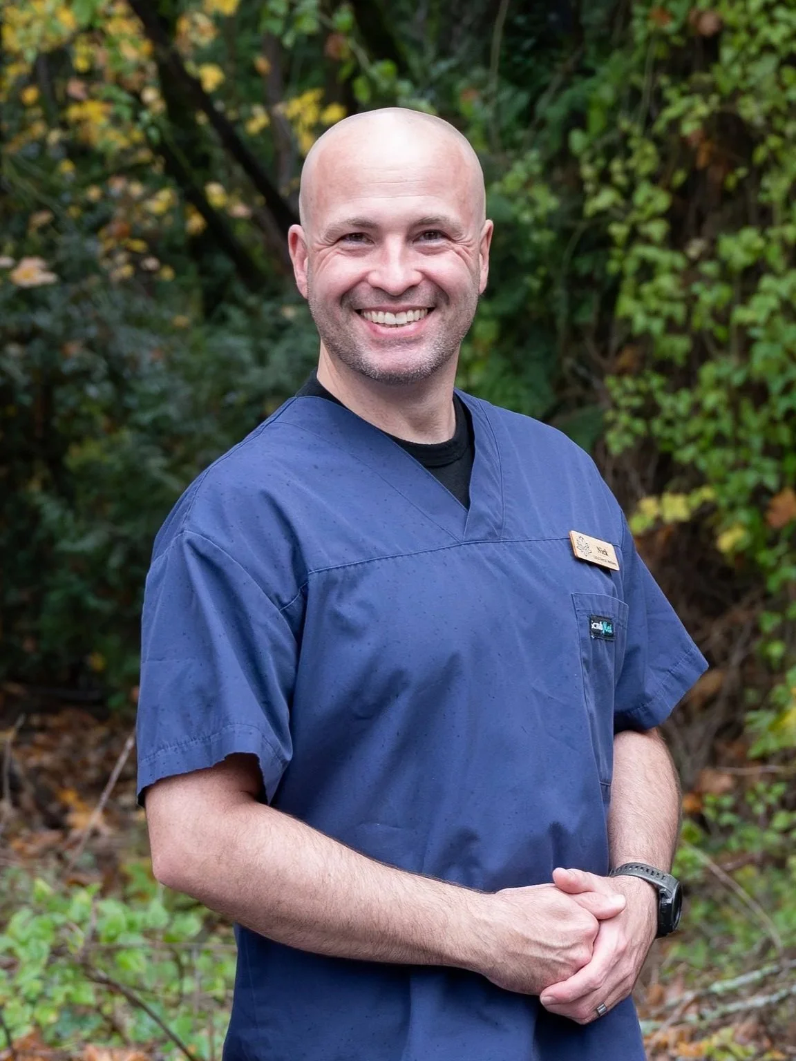A male healthcare professional, smiling, wearing blue scrubs and a black watch, standing outdoors with green foliage in the background.