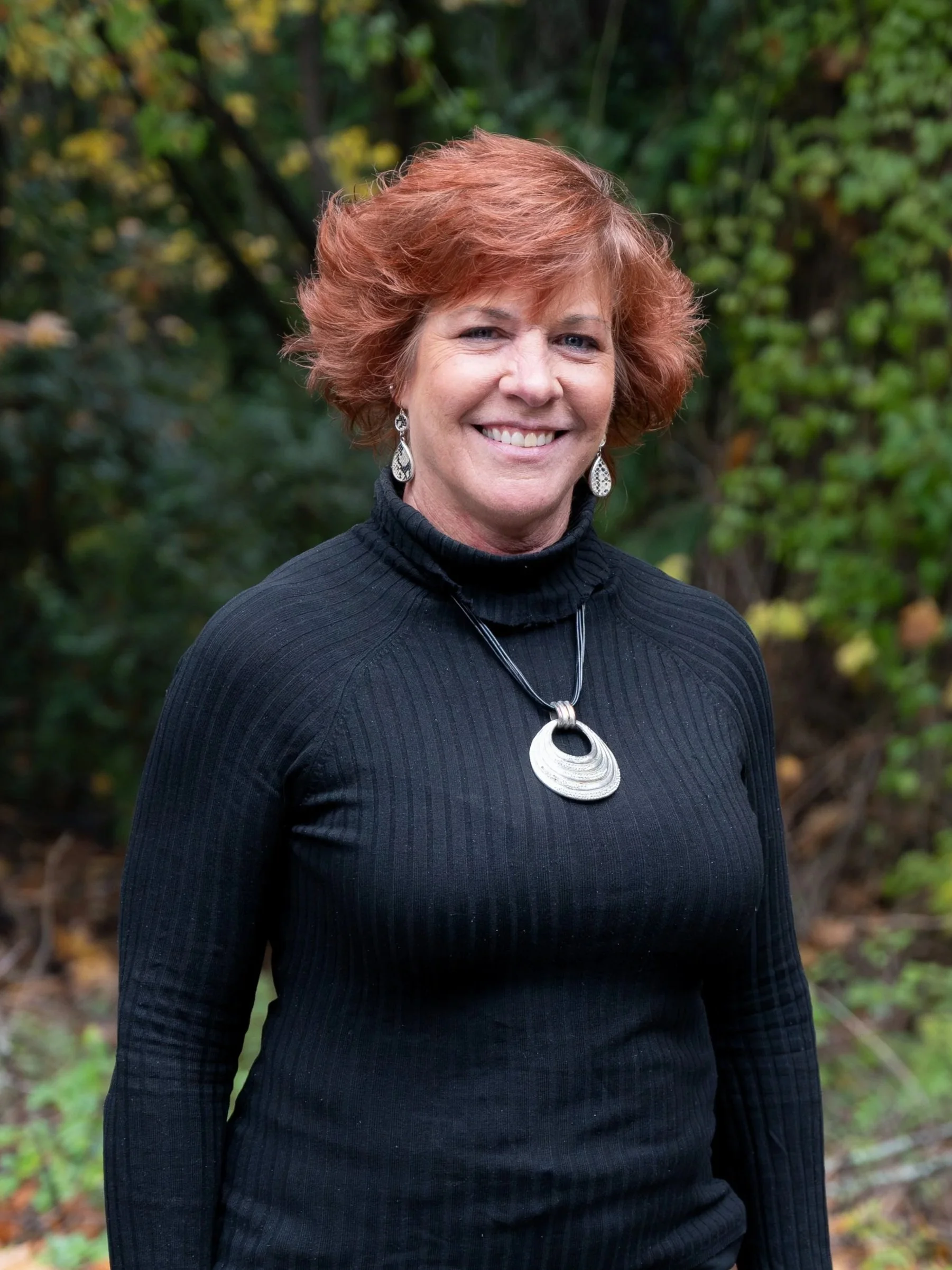 Smiling woman in a patterned sweater standing outdoors with trees in the background.