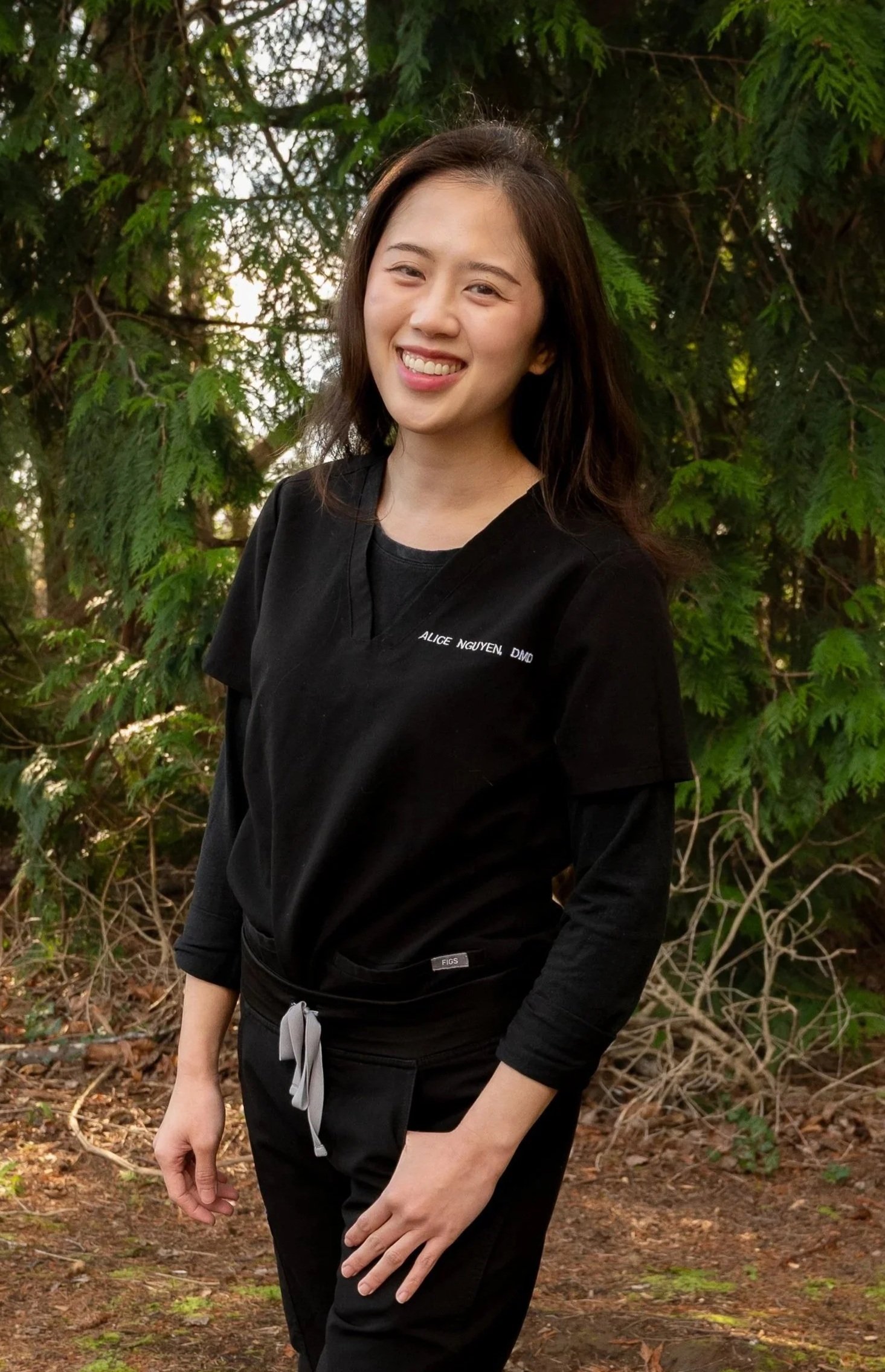 A young woman smiling outdoors, wearing black scrubs with her name embroidered on the chest, standing in front of green leafy trees.