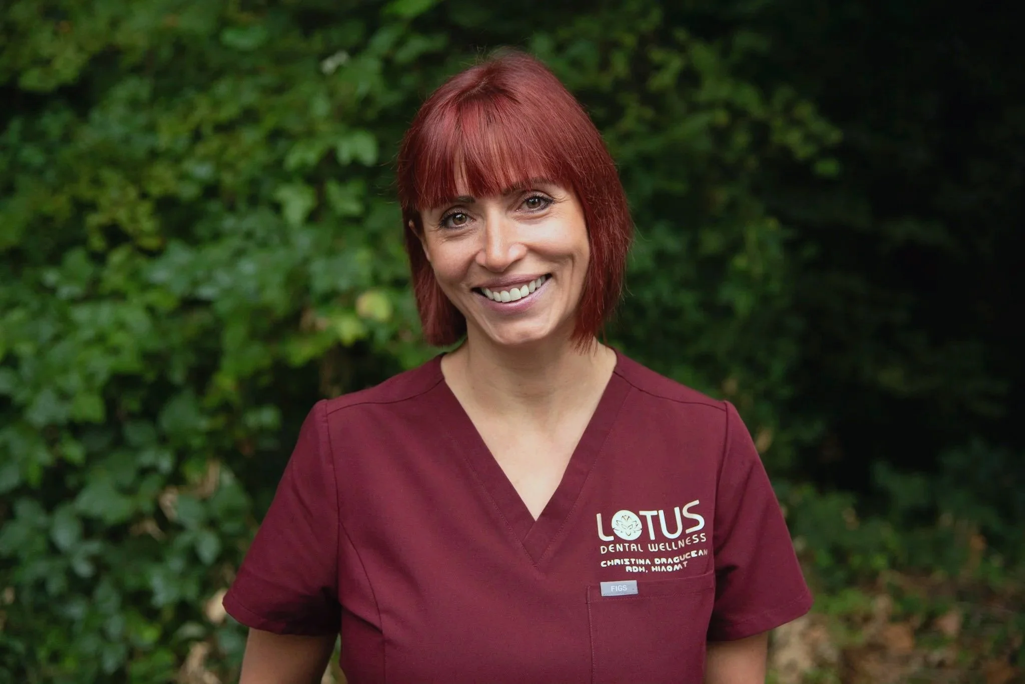 Cristina in a maroon dental uniform with "Lotus Dental Wellness" logo, standing outdoors with greenery background.