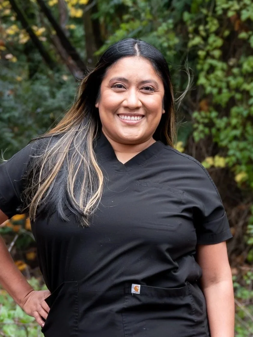 A person with long dark hair wearing a black shirt smiling in front of green foliage outdoors.