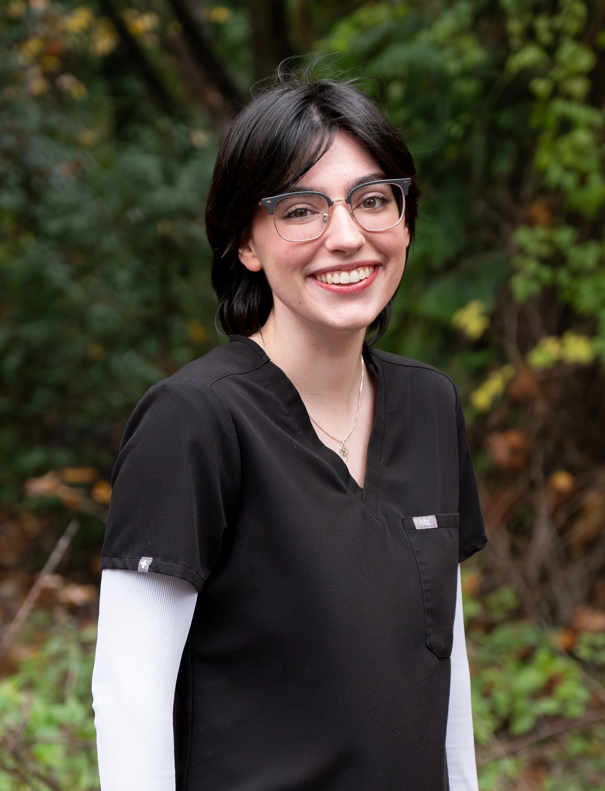 A young woman with long brown hair wearing a blue scrubs top, smiling and standing outdoors in front of dense bushes and vines.