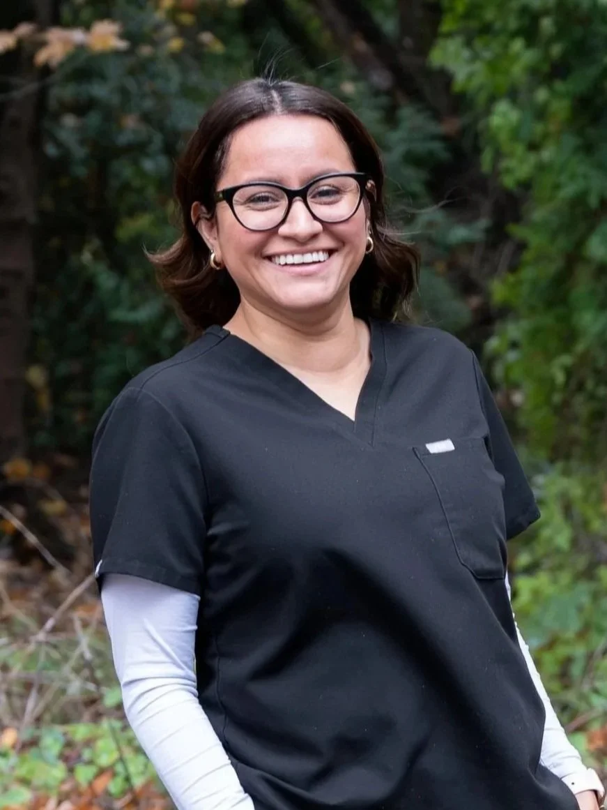 A dental assistan with brown hair, glasses, and a black scrub top standing outdoors in a wooded area, smiling.