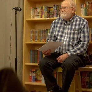 Older man with glasses and a beard, wearing a plaid shirt, sitting on a chair in front of a bookshelf, holding papers, in a library or bookstore setting.