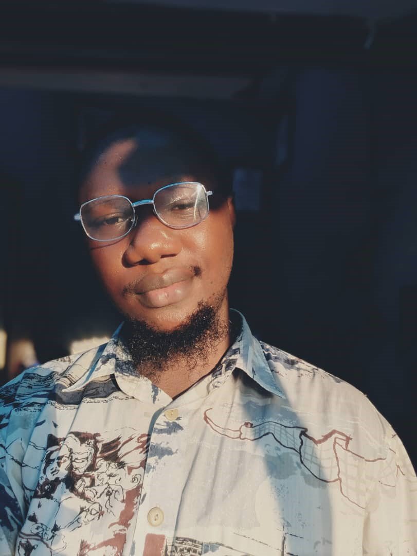A young man with glasses and a beard wearing a patterned button-up shirt, smiling in sunlight with a dark background.