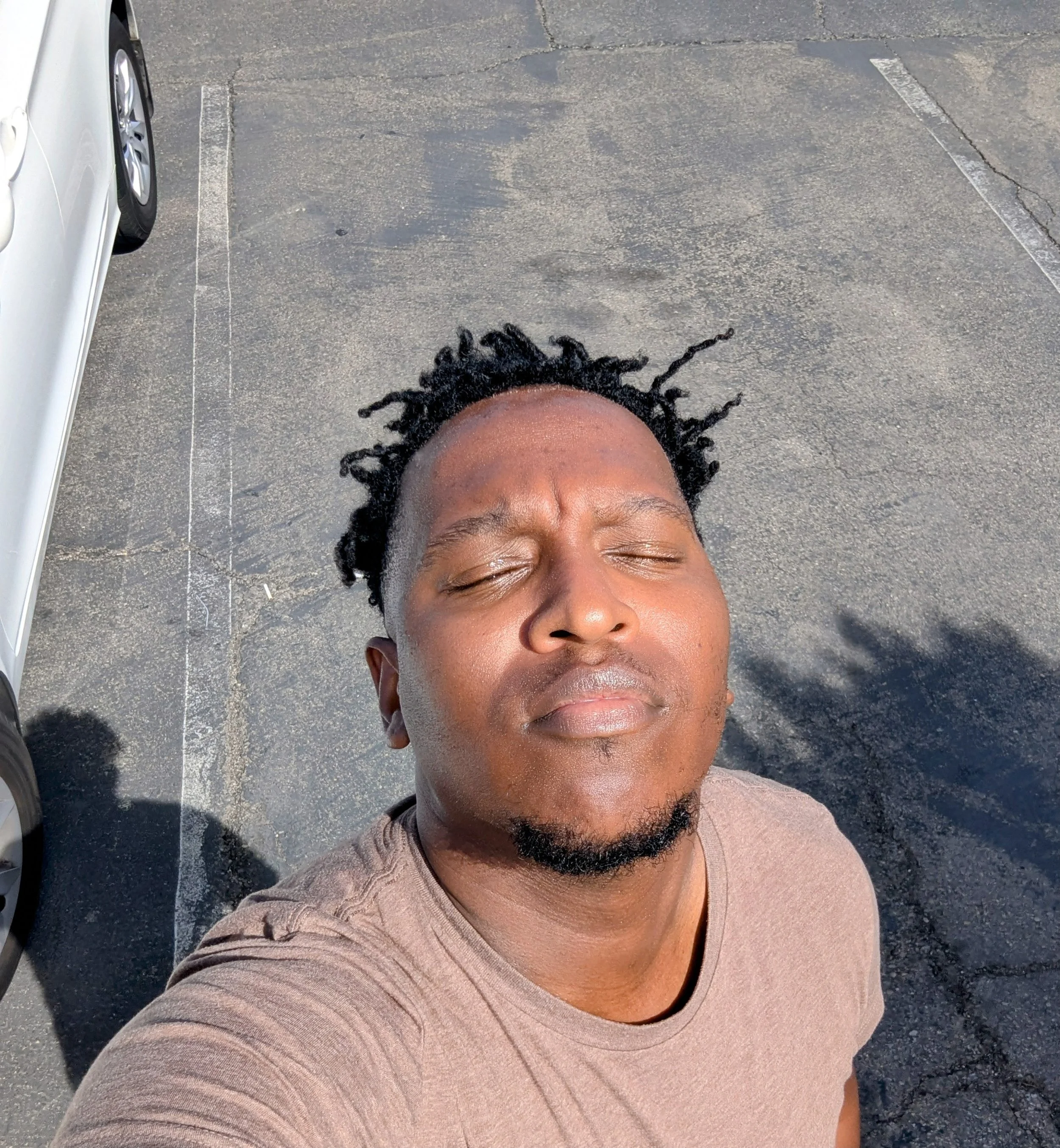 A person with closed eyes and curly black hair taking a selfie on a parking lot in bright sunlight.