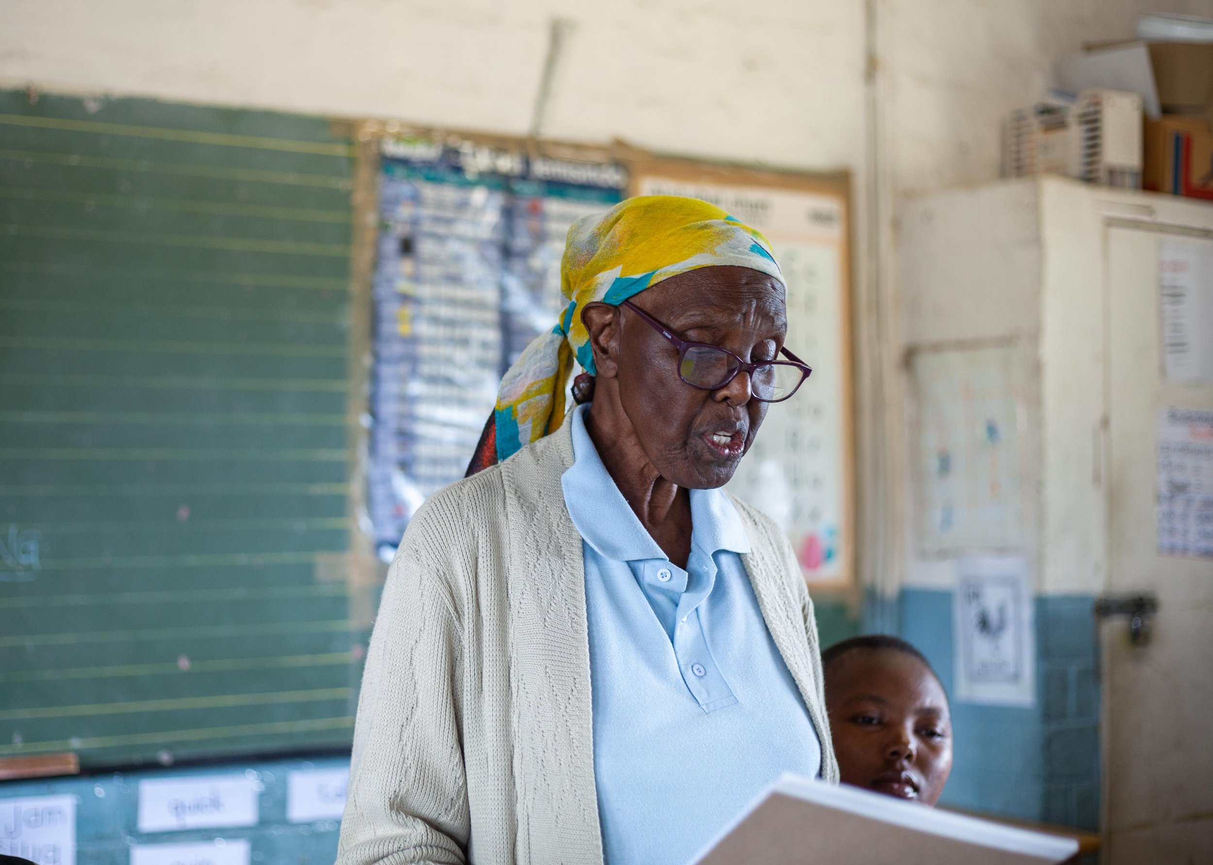 elder standing in blue polo, yellow patterned head scarf, and glasses in front of a schoolroom blackboard