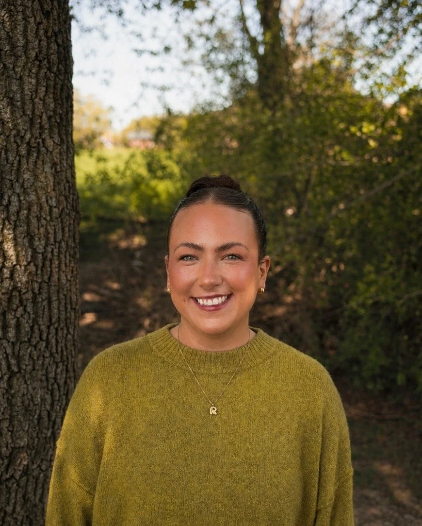 A woman with a bun hairstyle smiling outdoors in front of a tree and greenery in the background.