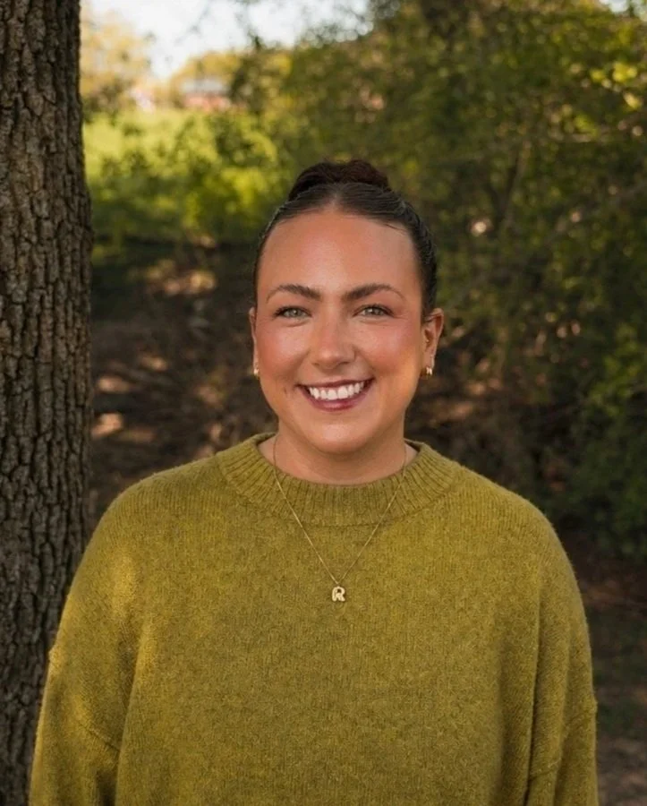 A smiling woman standing outdoors near a tree in a park with green and yellow foliage in the background, wearing a green sweater and gold jewelry.