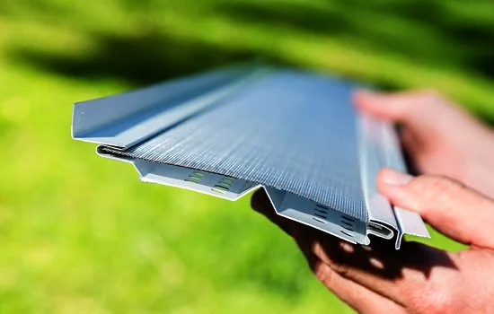 Hands holding a piece of metal gutter guard outdoors on a sunny day.