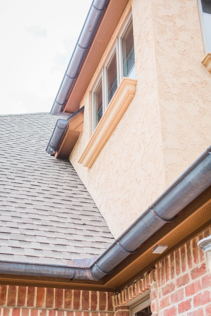 Close-up of a house exterior featuring a beige stucco wall, asphalt shingle roof, and brown gutters. The image shows a section of windows and part of a brick facade.