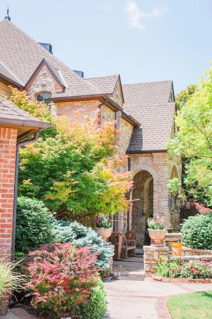 Stone house with steep roof, surrounded by colorful garden and foliage.
