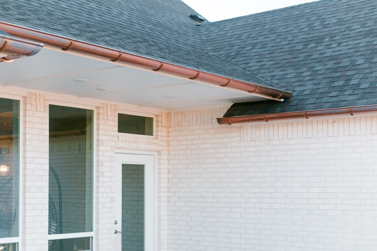 Exterior view of a house corner with white brick walls, large windows, a white door, shingled roof, and copper gutters.