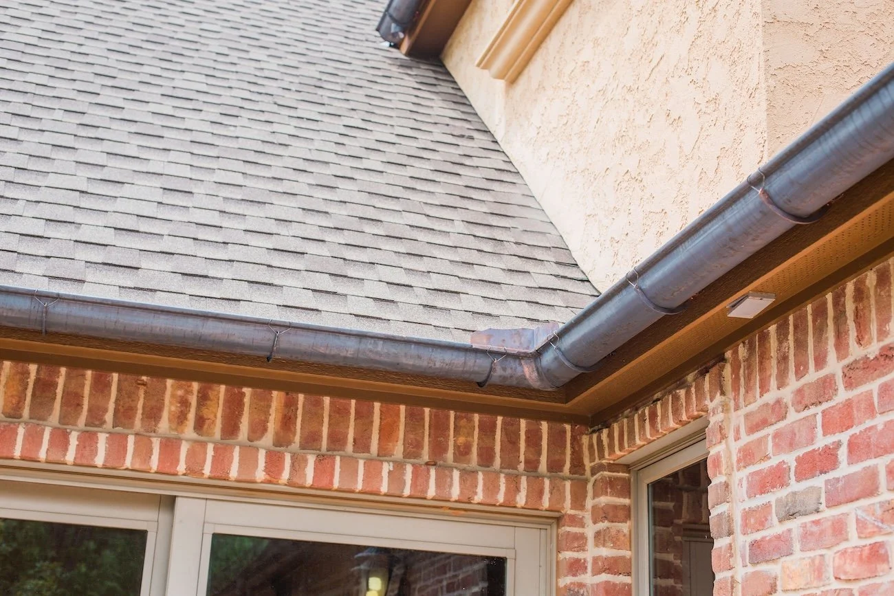 Close-up of a brick house corner with gray asphalt shingle roofing and dark metal gutters.