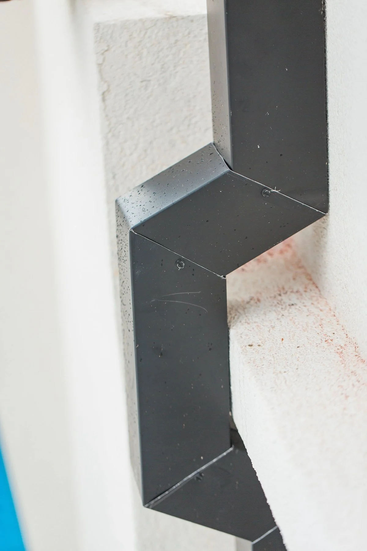 Close-up of a black metal downspout attached to a white stucco wall with visible corner joints.