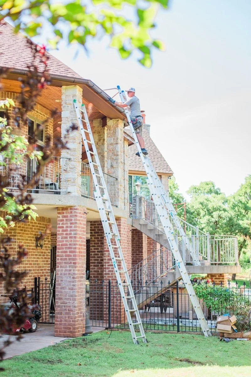 Person working on a ladder fixing a gutter on a brick house with a patio and greenery.