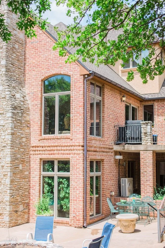 Brick house exterior with stone chimney, large windows, and a balcony. Outdoor patio with chairs, a small table, and potted plants. Green foliage visible in the background.