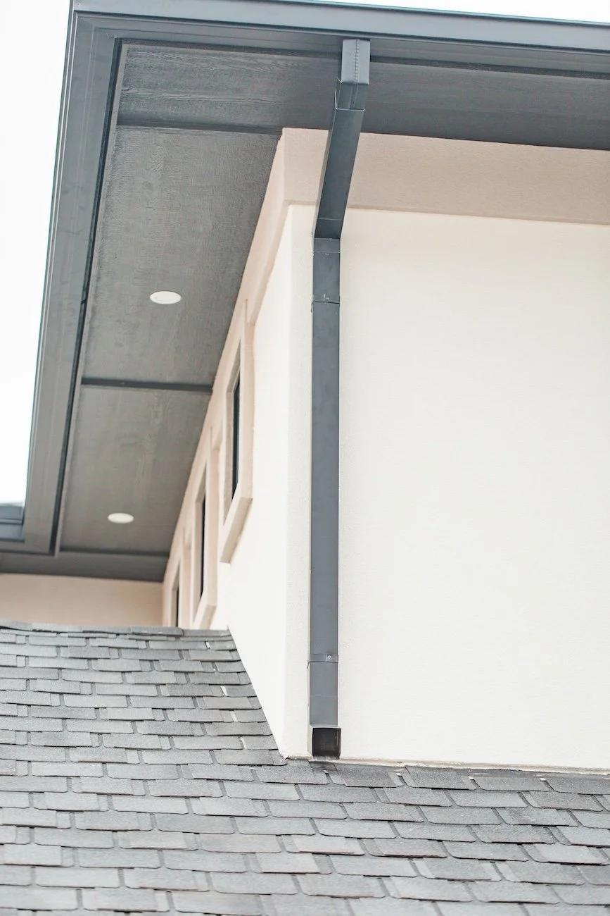 Close-up of a house roof corner showing shingles, soffit with recessed lights, and a dark downspout against a light exterior wall.