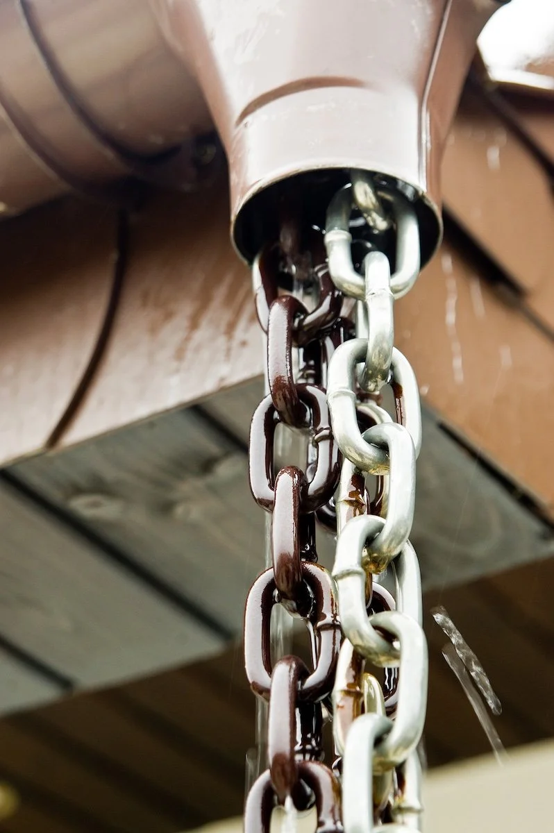 Close-up of a rain chain attached to a brown gutter, with silver and brown chain links.