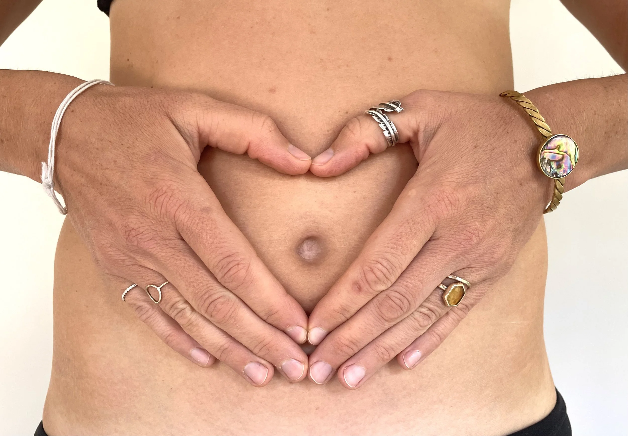 Person making a heart shape with their hands over their bare stomach, wearing rings and bracelets.